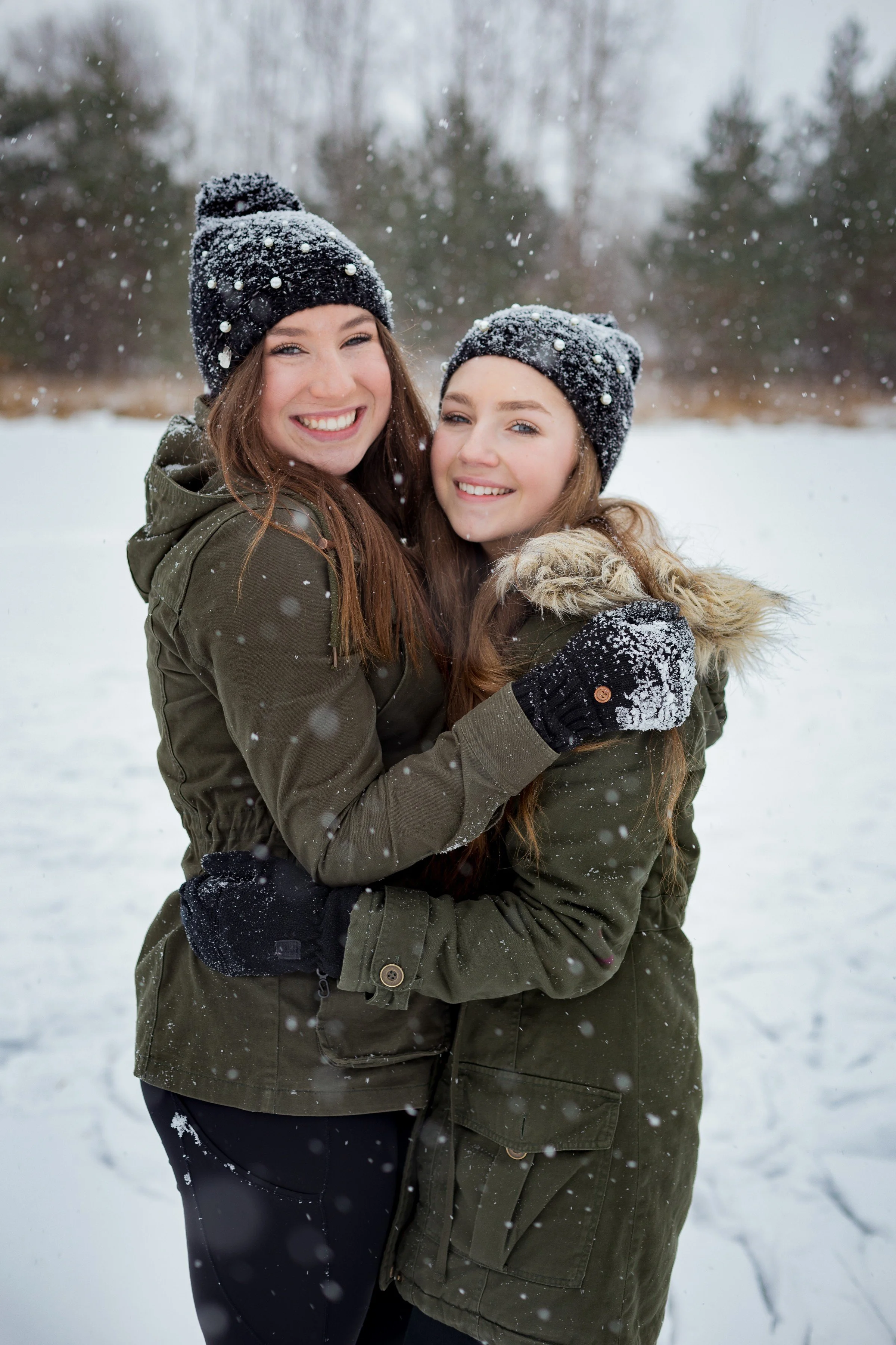 Two young women in winter coats and beanies hugging in a snowy outdoor setting, smiling at the camera.