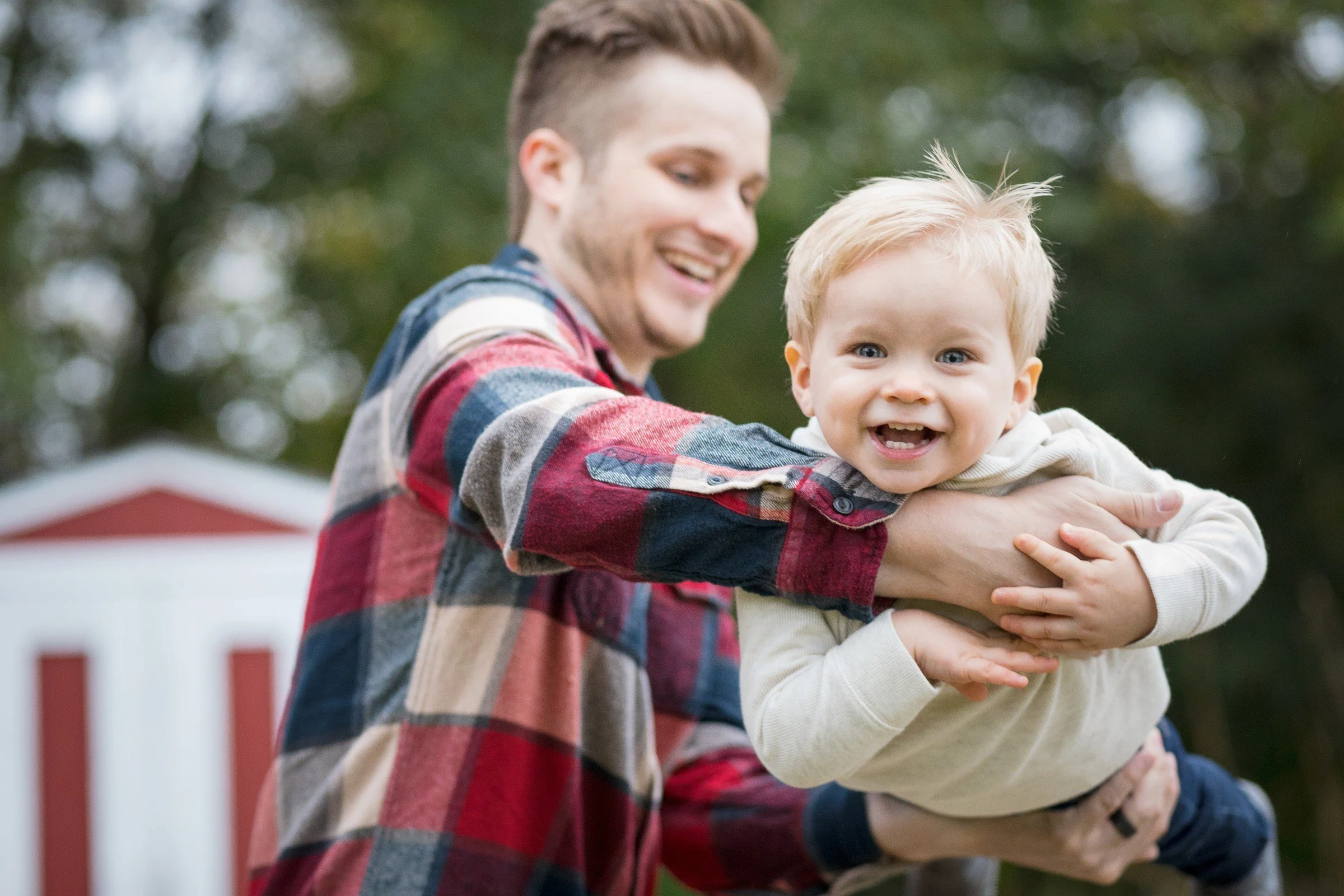 A man holding a smiling young boy outdoors, with a blurred house and trees in the background.