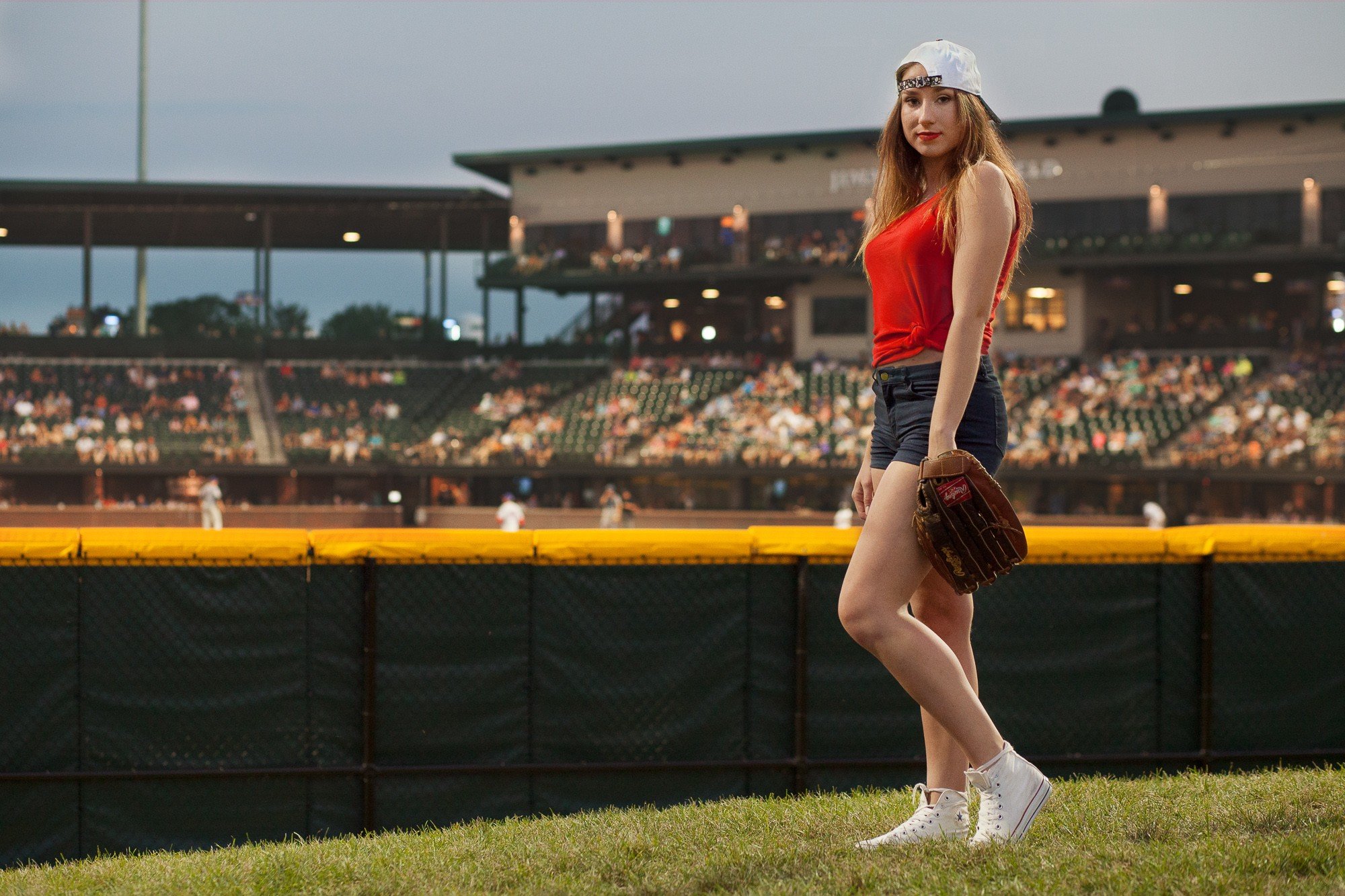 A young woman stands on the baseball field in a stadium, wearing a red tank top, denim shorts, white sneakers, and a baseball glove, with the crowd in the background.