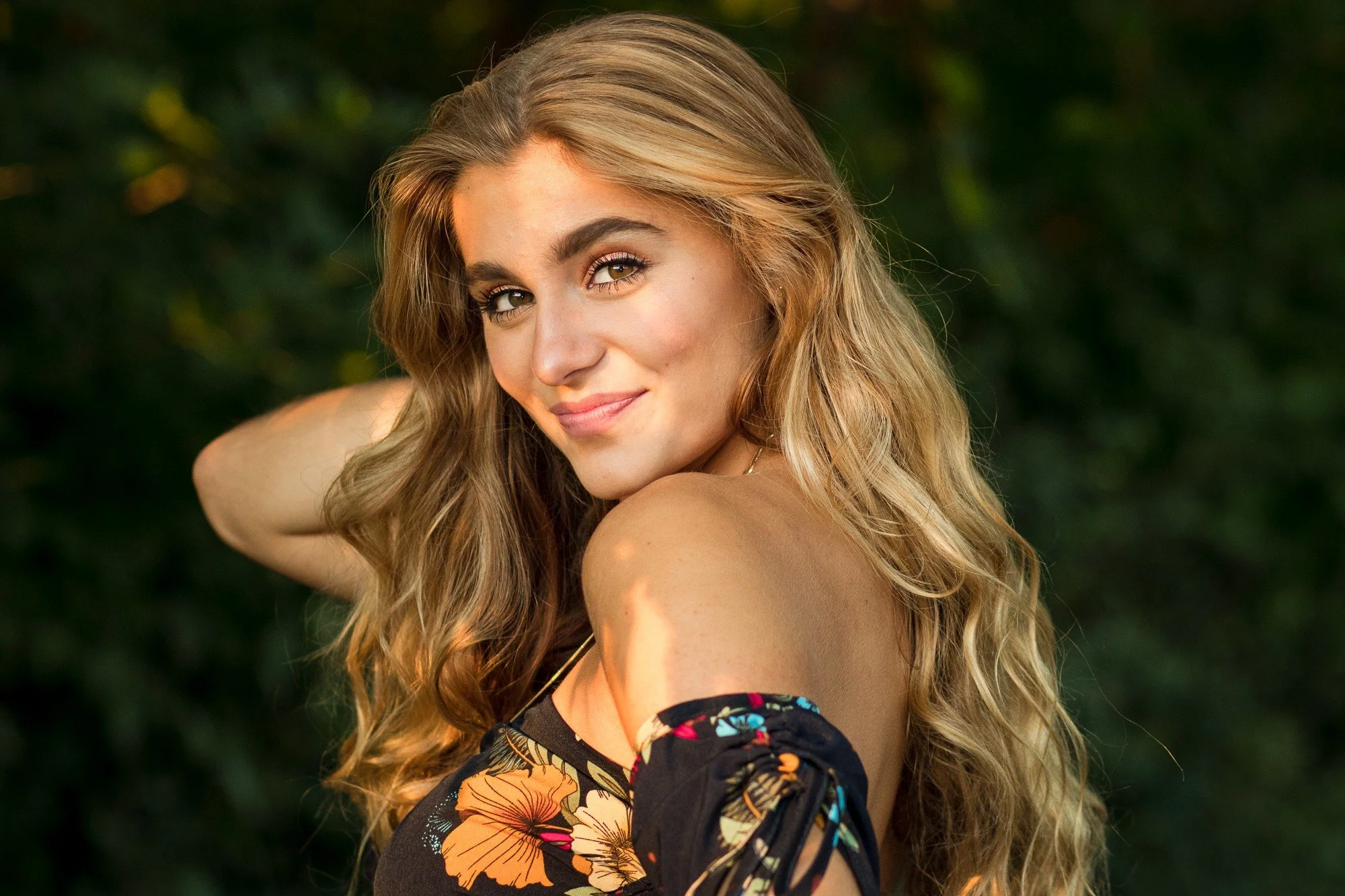 Young woman with long, wavy blonde hair smiling at camera, posing outdoors with greenery in the background.