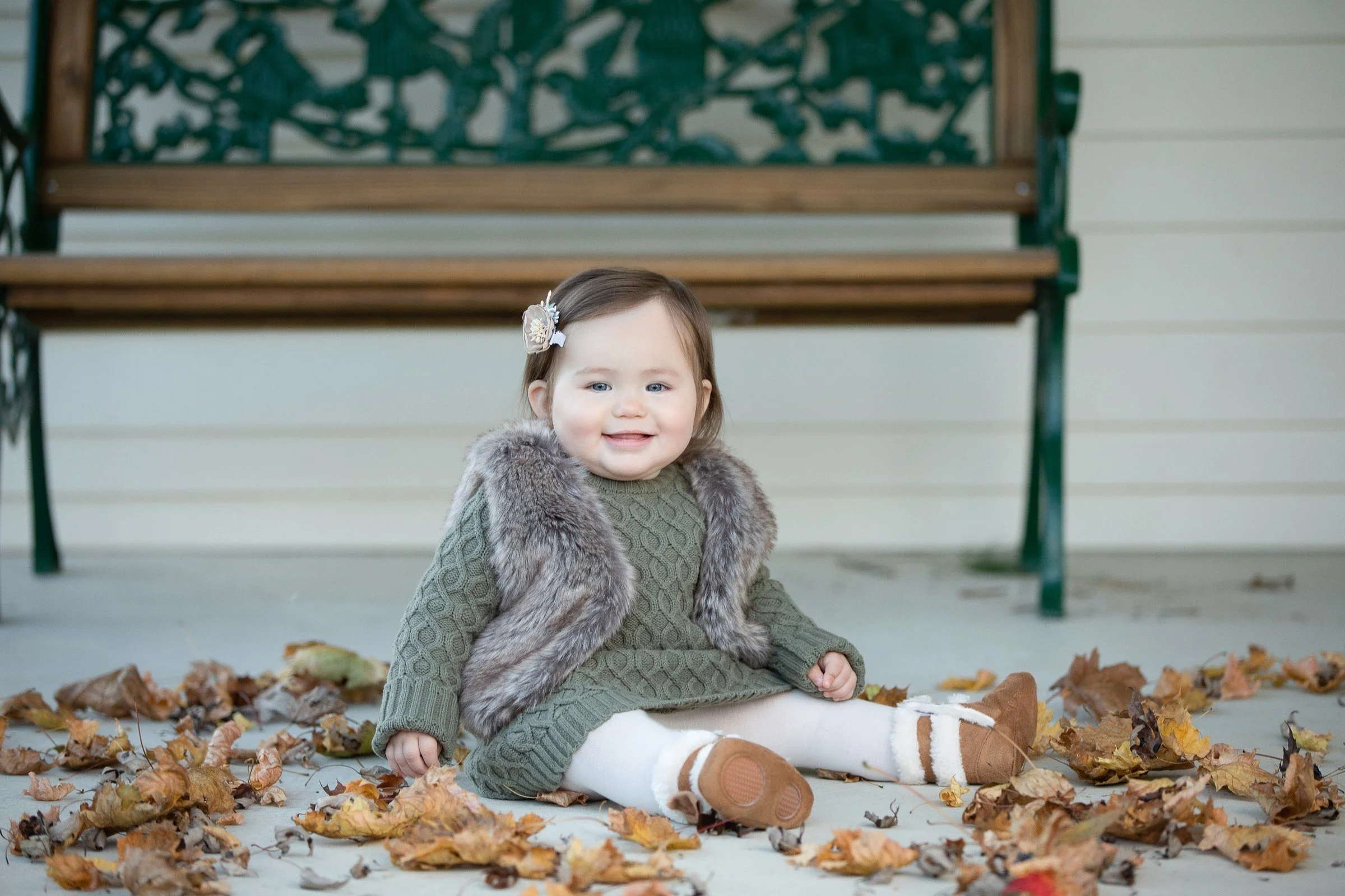 A young girl sitting on the ground surrounded by fallen autumn leaves, smiling, with a bench behind her.