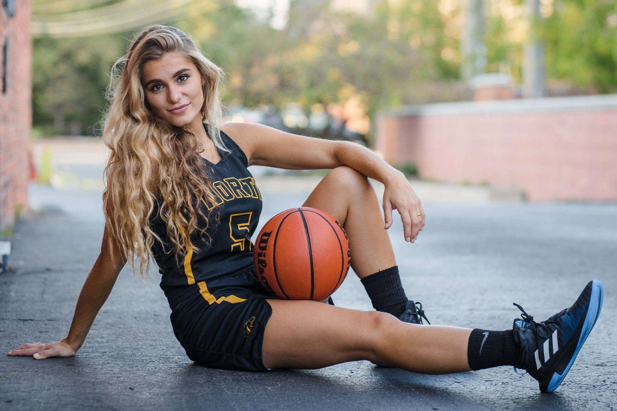 A young woman with long, wavy blonde hair, wearing a black basketball uniform, sitting on an outdoor basketball court with a basketball resting on her lap, smiling at the camera with trees and a brick wall in the background.