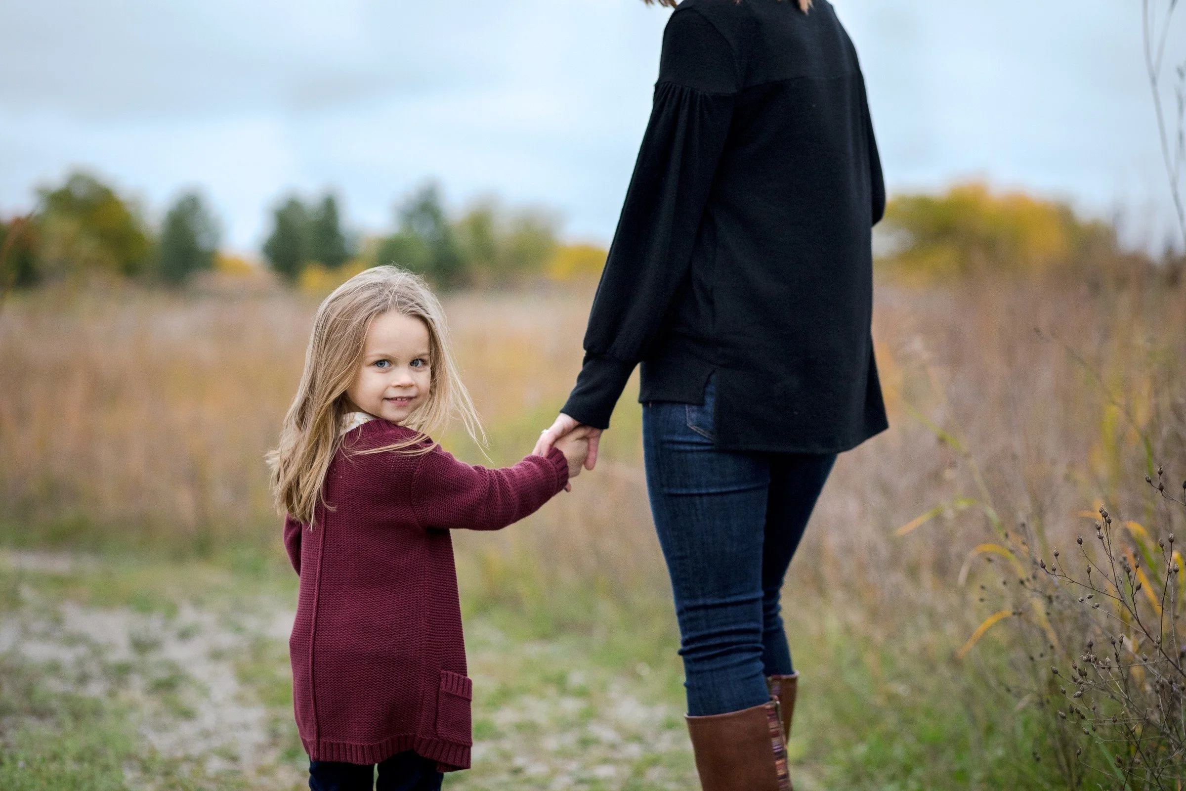 A young girl with long blonde hair and a burgundy sweater holding hands with an adult woman dressed in dark clothing, walking outdoors in a field with trees in the background during autumn.
