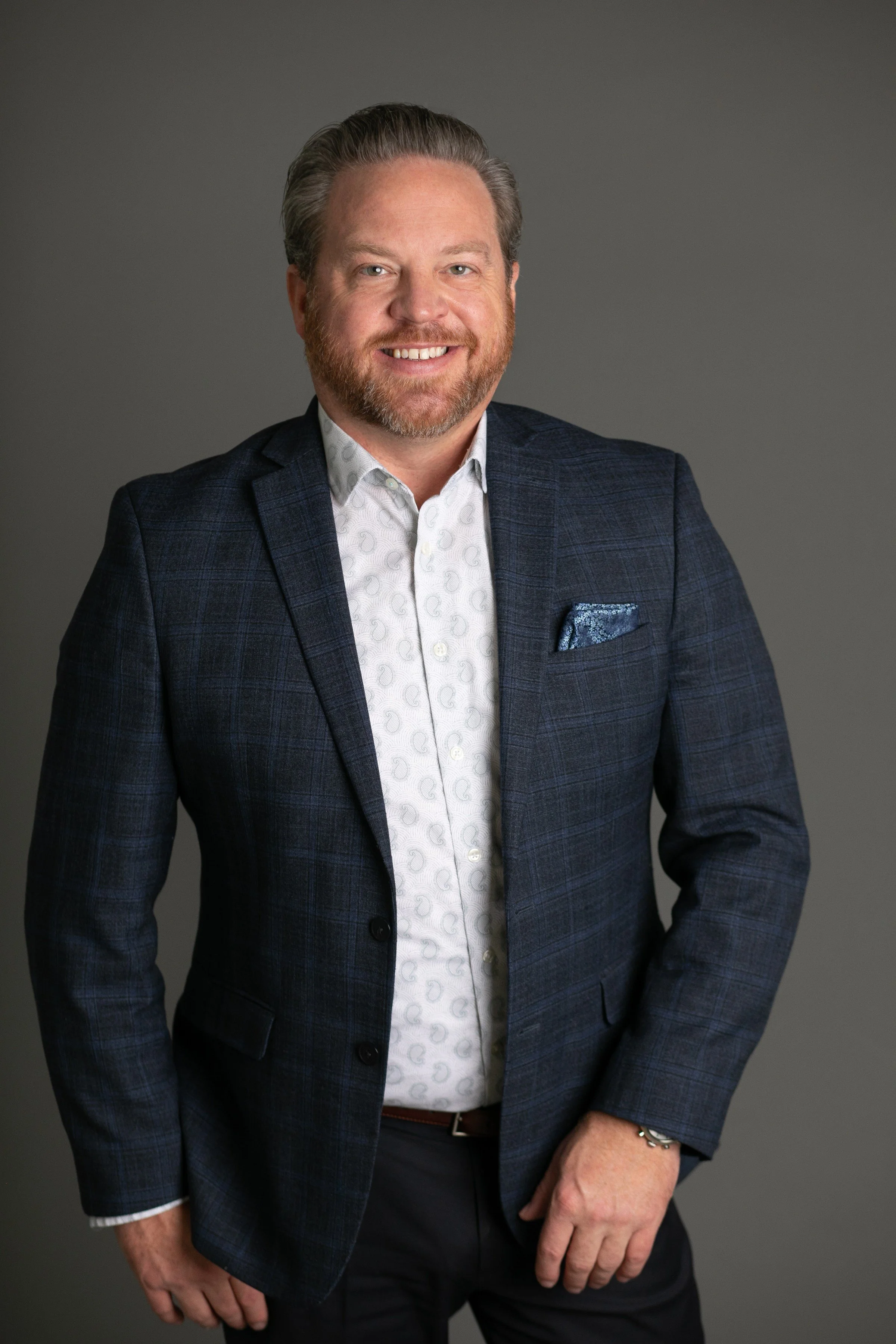 A man with light skin, short, light brown hair, and a beard, smiling, wearing a dark blue plaid blazer, a white patterned dress shirt, and black pants, standing against a gray background.