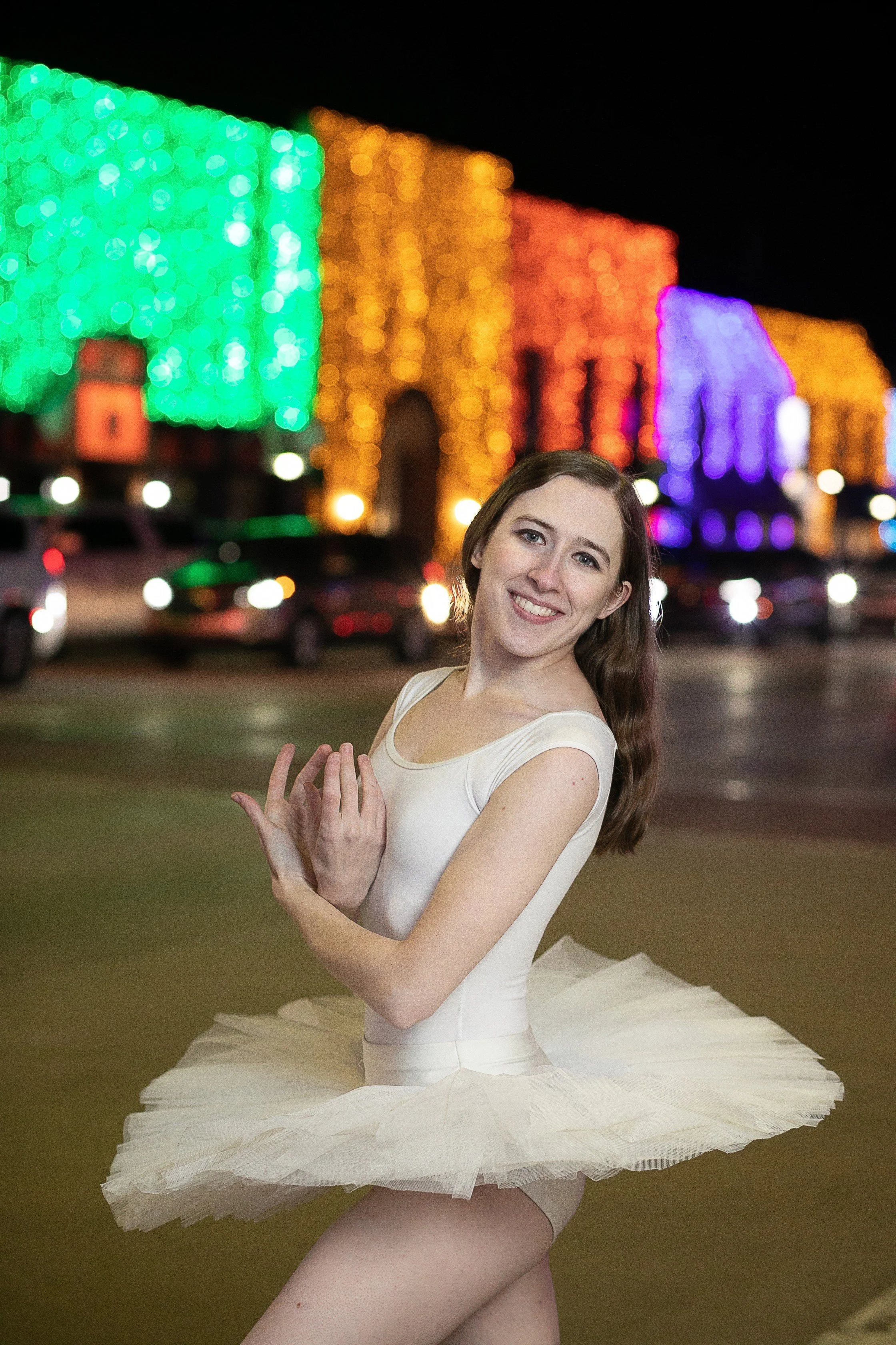 A ballet dancer in a tutu posing outdoors at night with colorful building lights in the background.