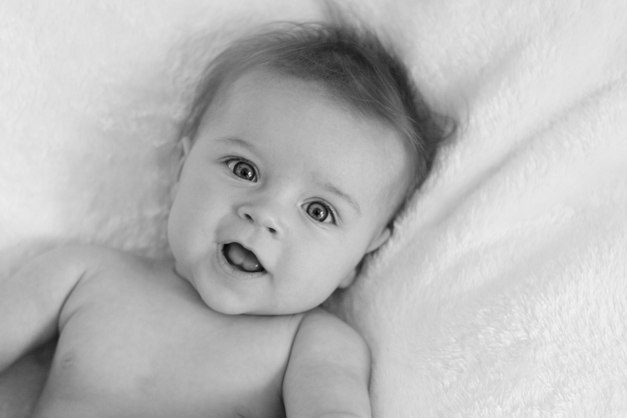 Black and white photo of a baby lying on a soft, fluffy surface, looking at the camera with curious eyes and mouth slightly open.