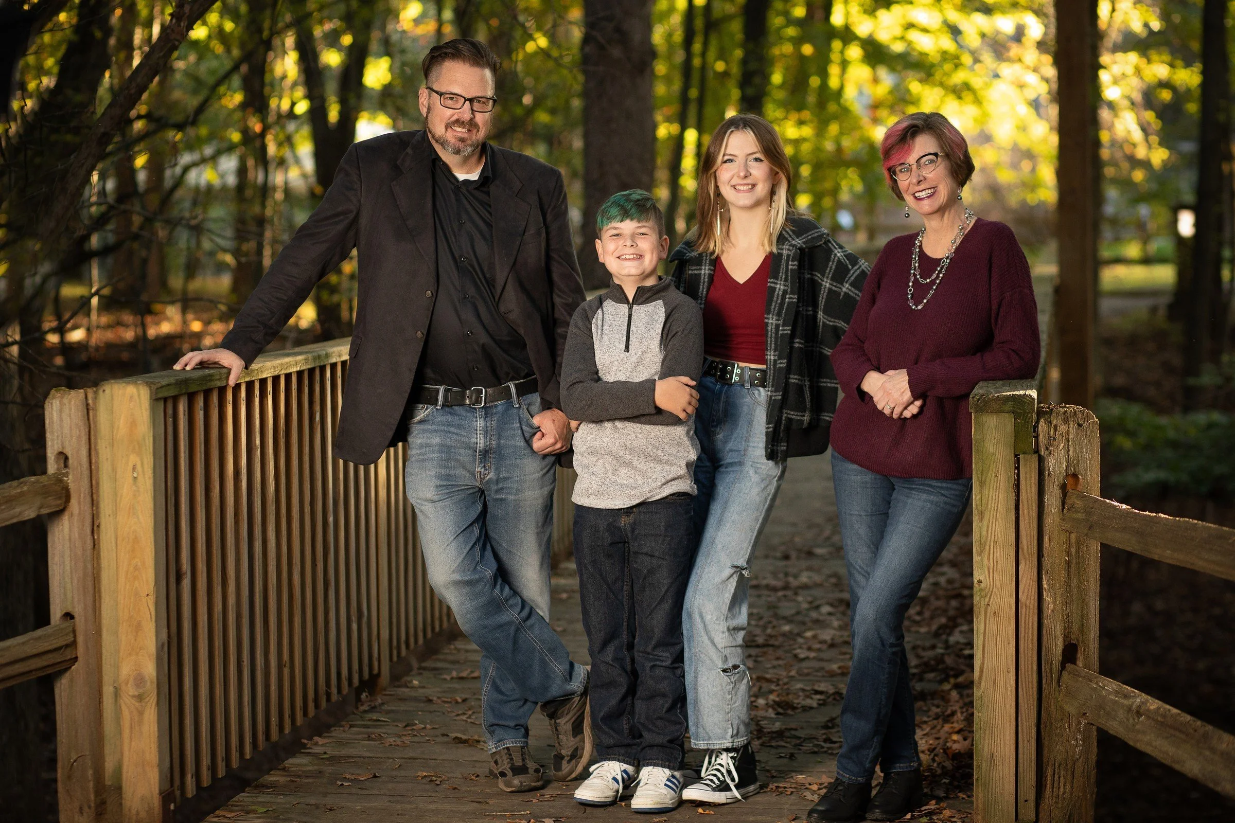 A family of five — two men, two women, and a boy — standing on a wooden bridge in a forest during autumn, smiling at the camera.