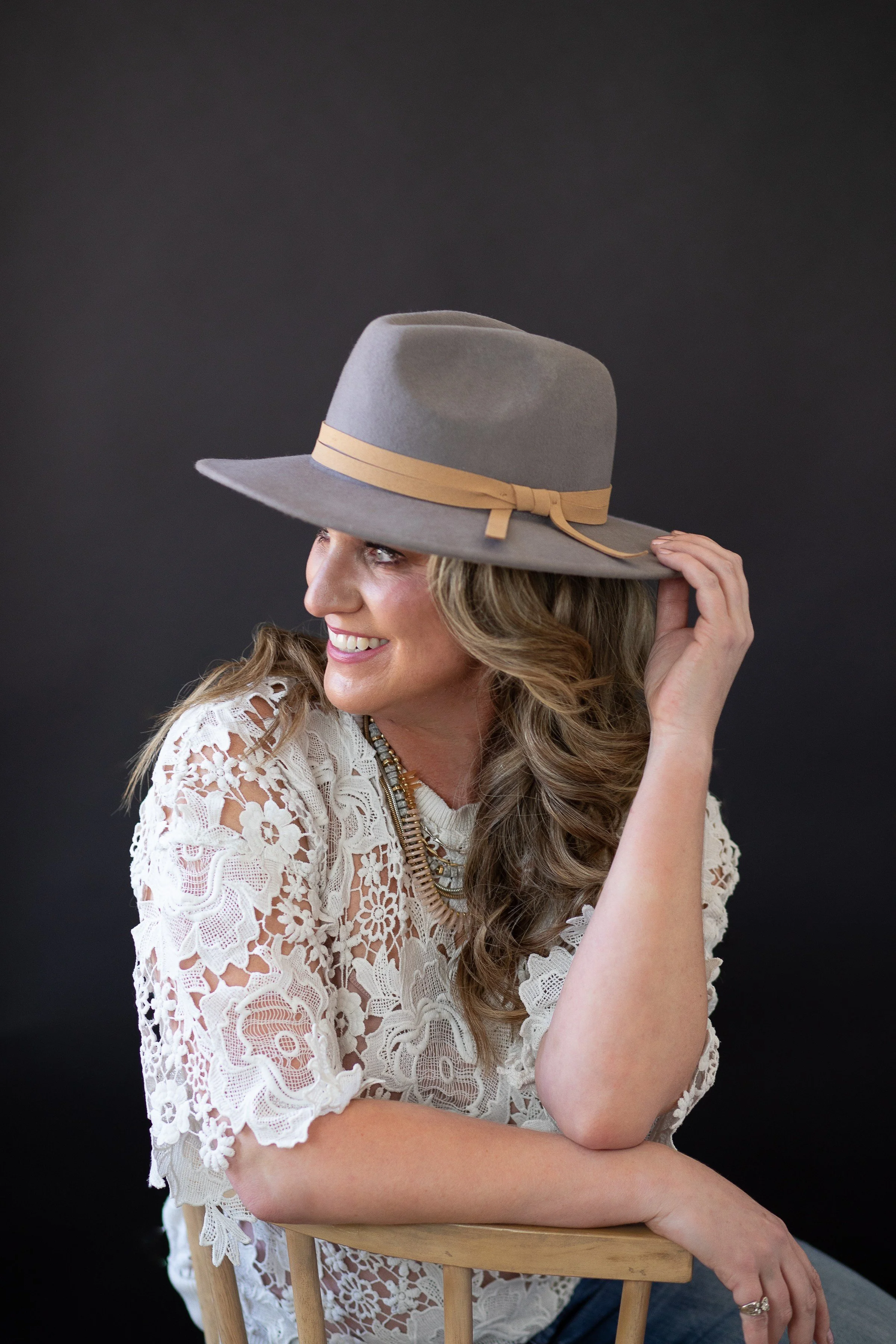 A woman smiling, wearing a wide-brimmed gray hat with a beige band, and a white lace top, sitting on a wooden chair against a black background.