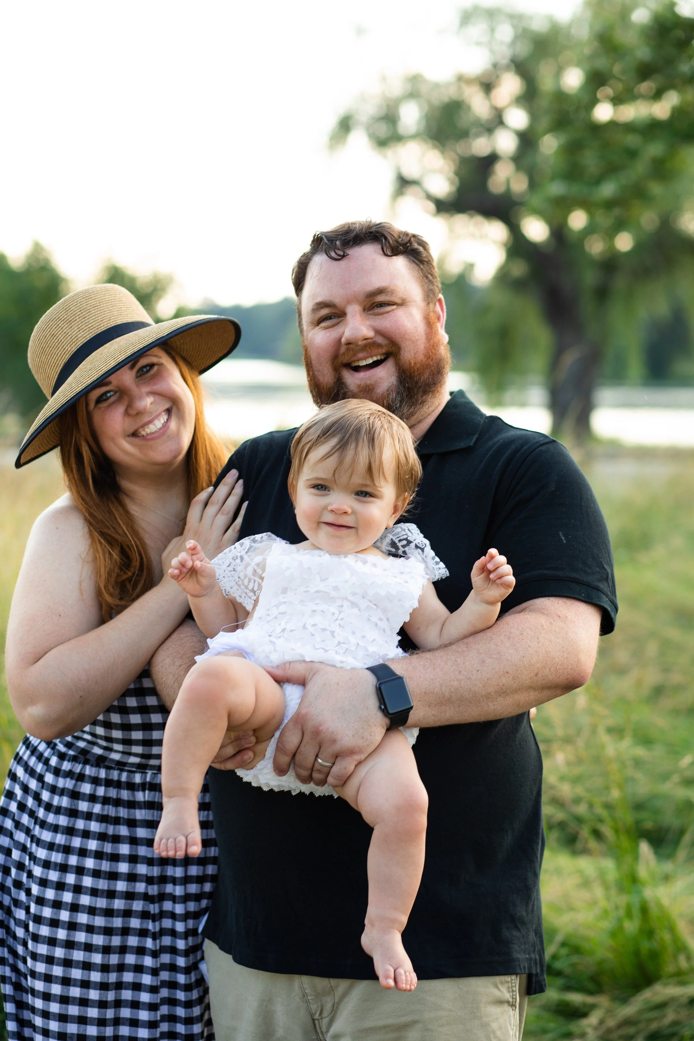 A happy family of three, a woman, a man, and a toddler girl, posing outdoors near a lake with trees in the background, in the evening light.