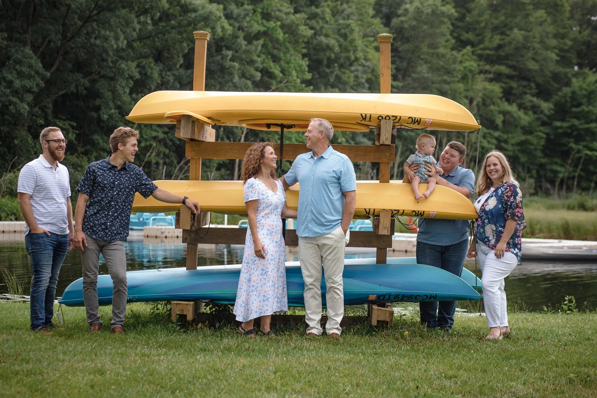 Group of seven people standing outdoors near a water, with a rack holding three kayaks or canoes, two yellow and one blue, while smiling and talking to each other.