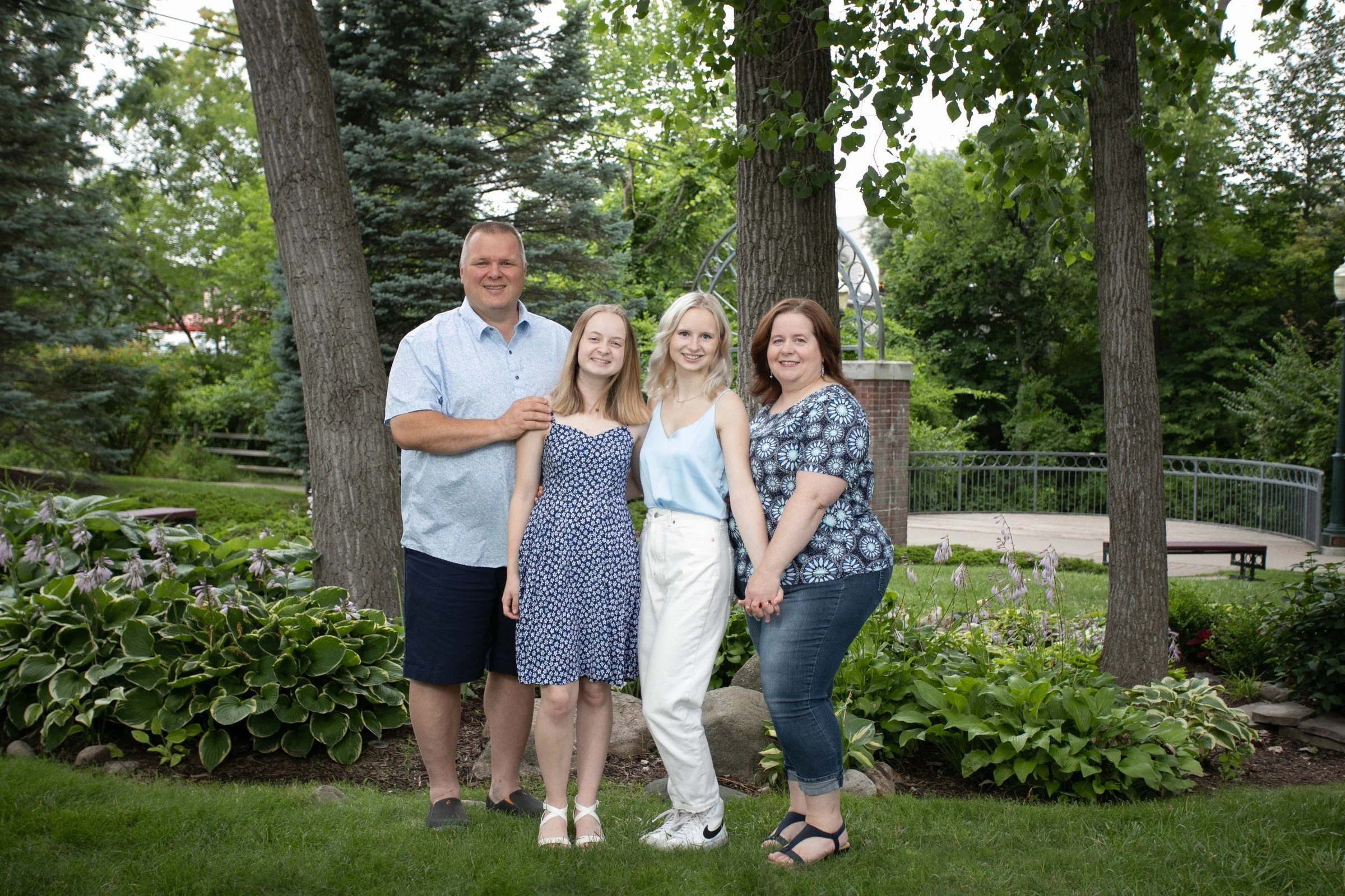 A family of four with two women and two teenagers, standing outdoors in a garden with trees, plants, and a bench in the background.