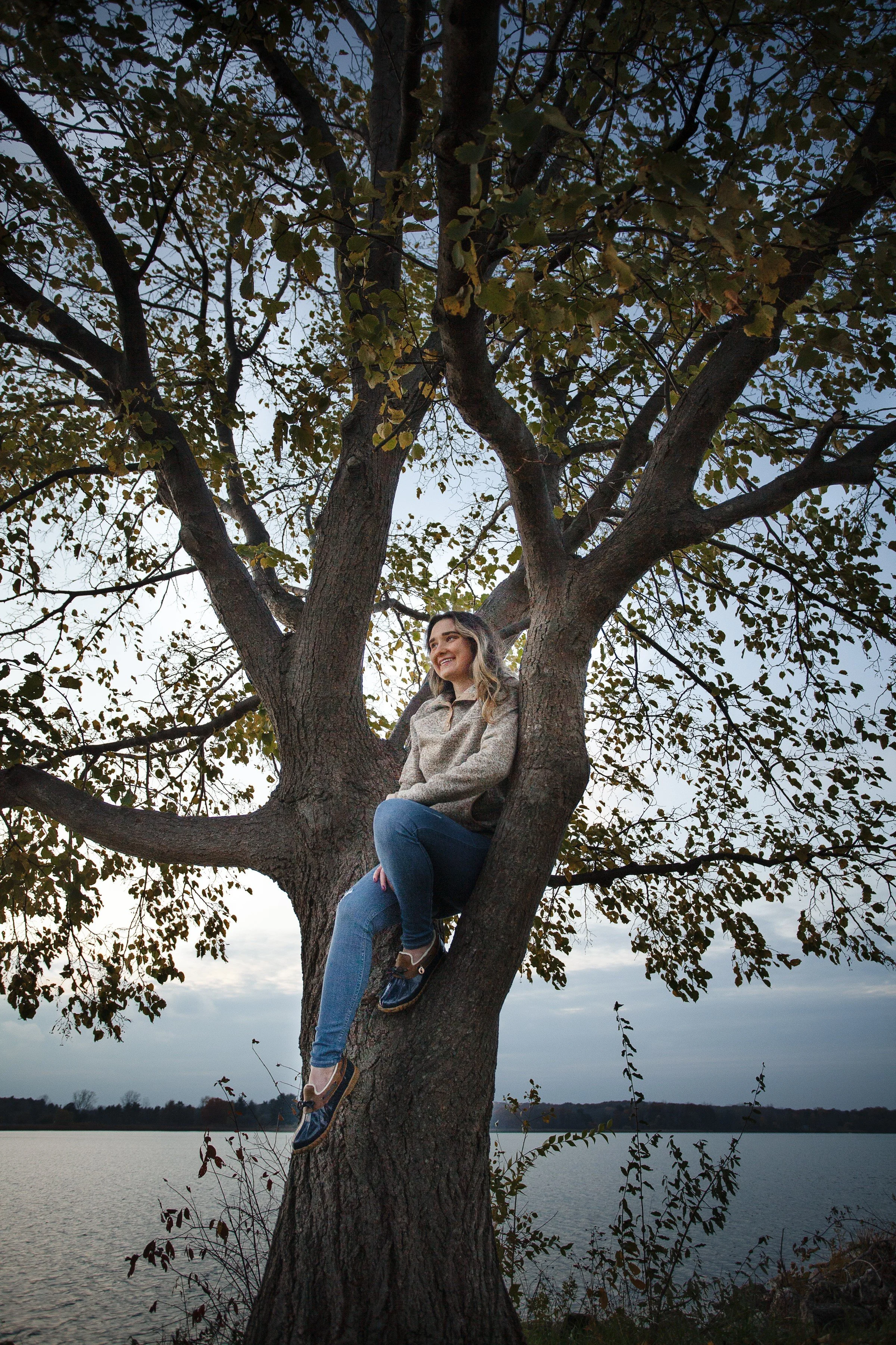 A young woman sitting on a tree branch near a lake at dusk, smiling and looking away from the camera.