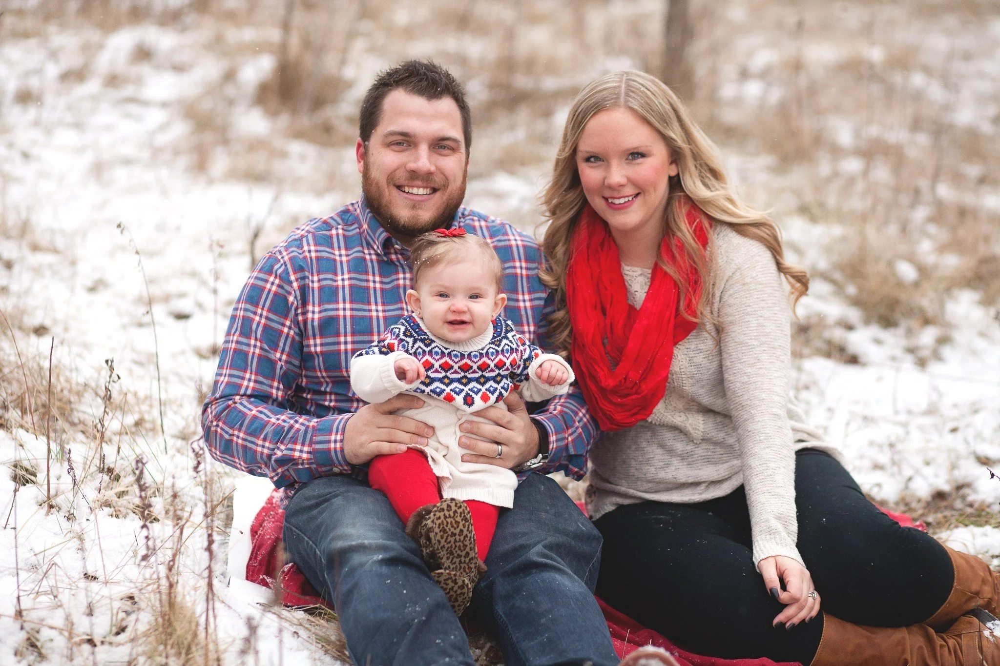 A family of three sitting outdoors in a snowy field, smiling at the camera. The father has a beard and is wearing a plaid shirt, holding a young girl in a festive sweater and red tights. The mother has long blonde hair, wearing a beige sweater and a 