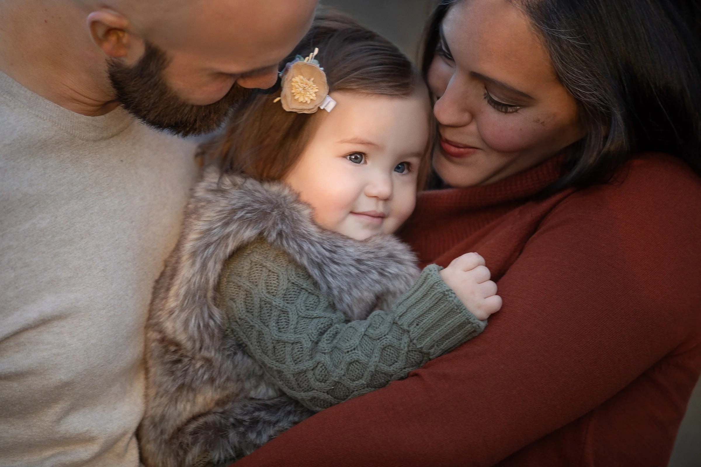 A family of three, with a man, a woman, and a young girl, closely hugging and sharing a tender moment outdoors.