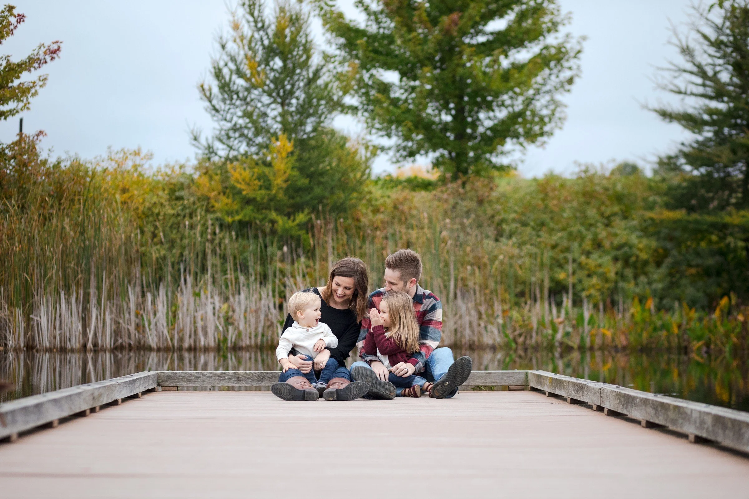 A family of four sitting on a wooden dock by a pond, surrounded by trees with fall foliage, enjoying a cozy moment together.