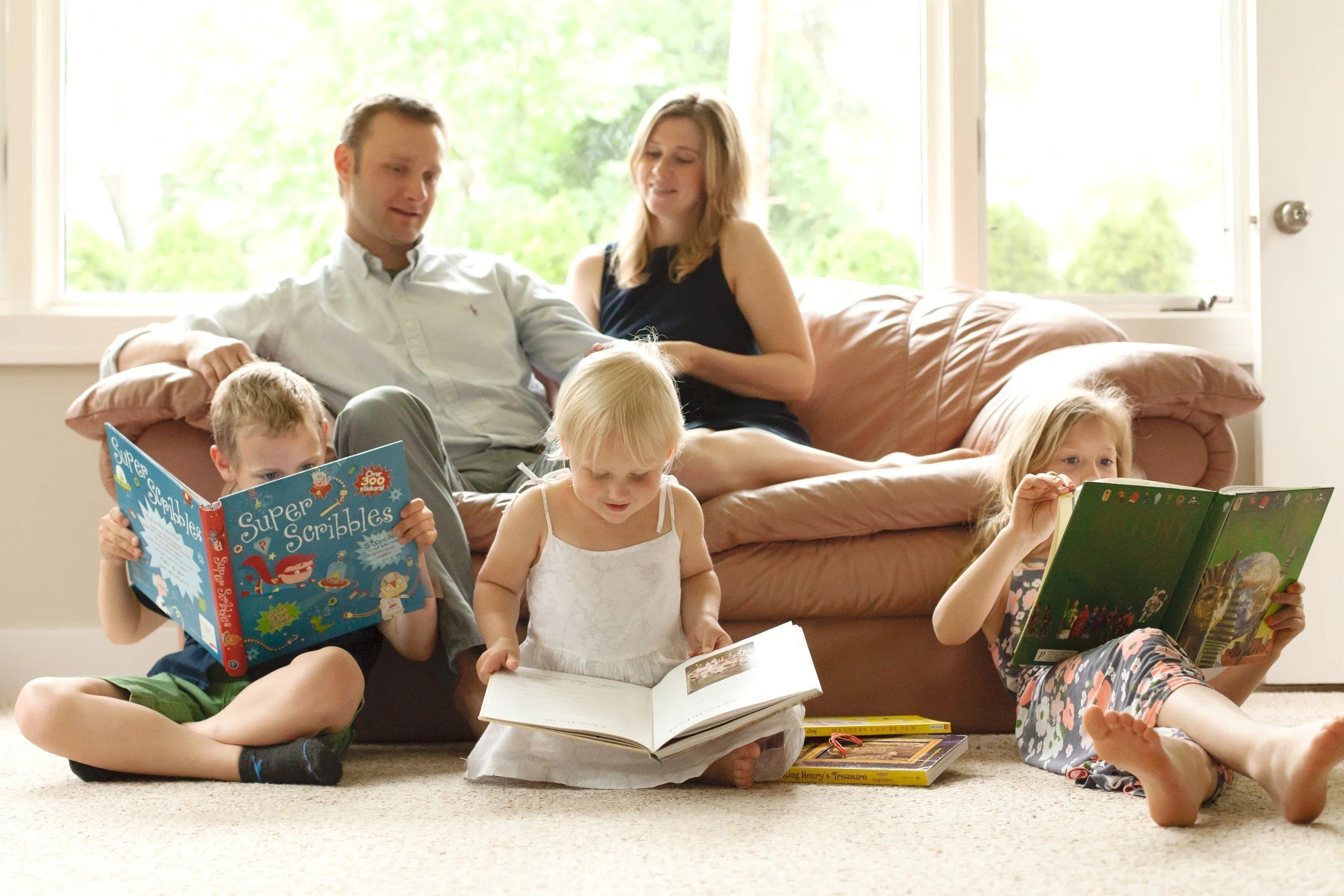 Family of five reading books in a living room with large windows and green trees outside.