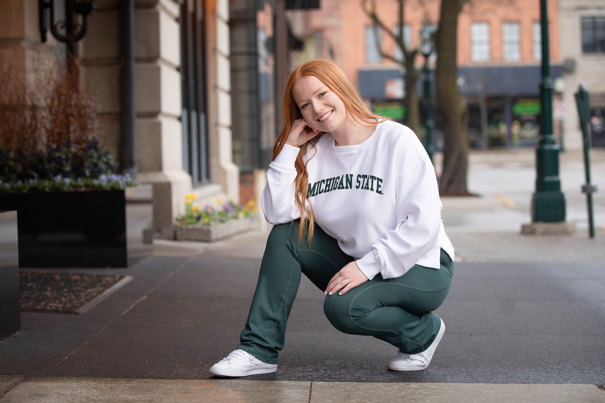A young woman with long red hair wearing a white Michigan State sweatshirt and dark green pants crouches on a city sidewalk, smiling at the camera.