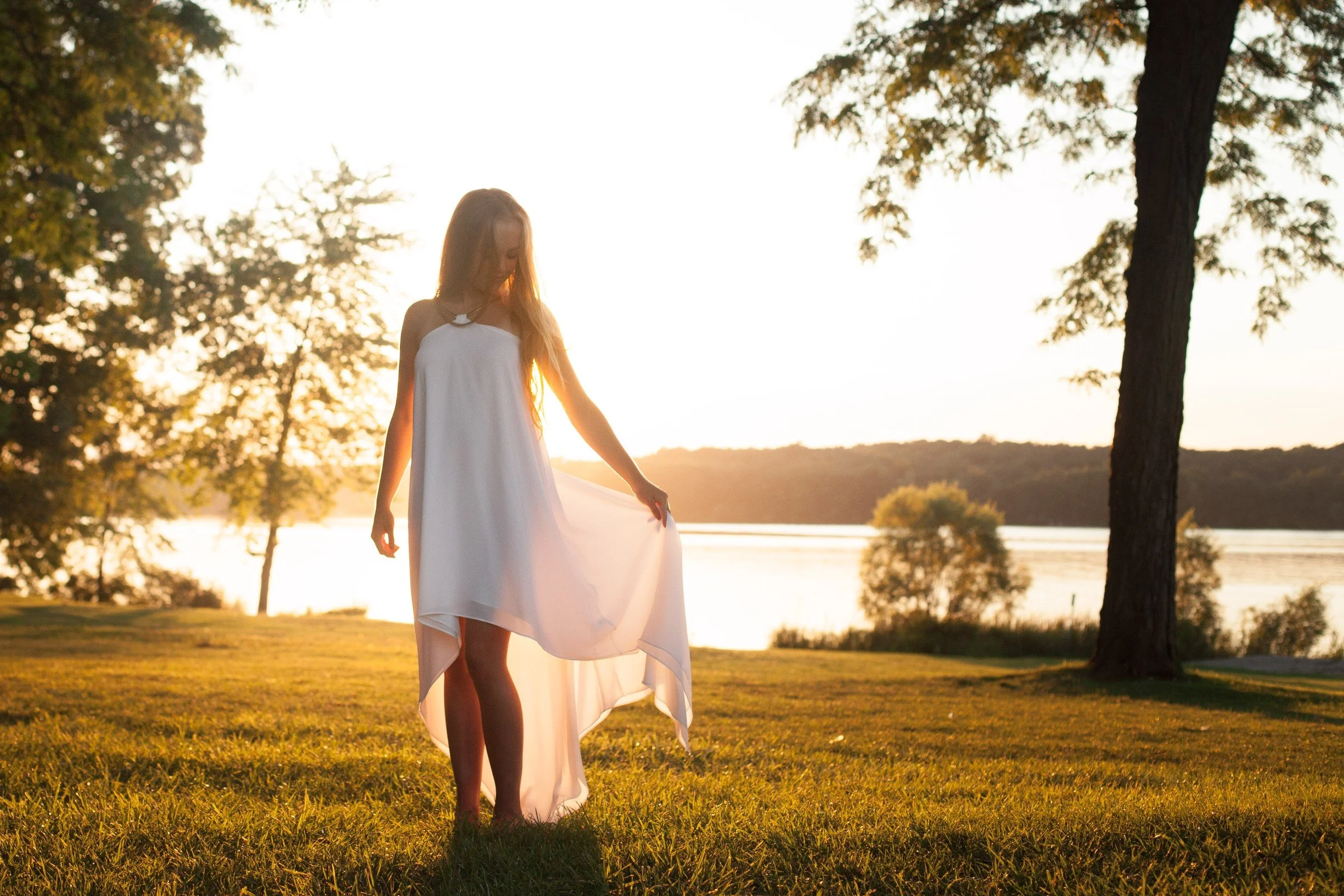 A woman in a white dress standing on grass near a body of water during sunset, with trees in the background.