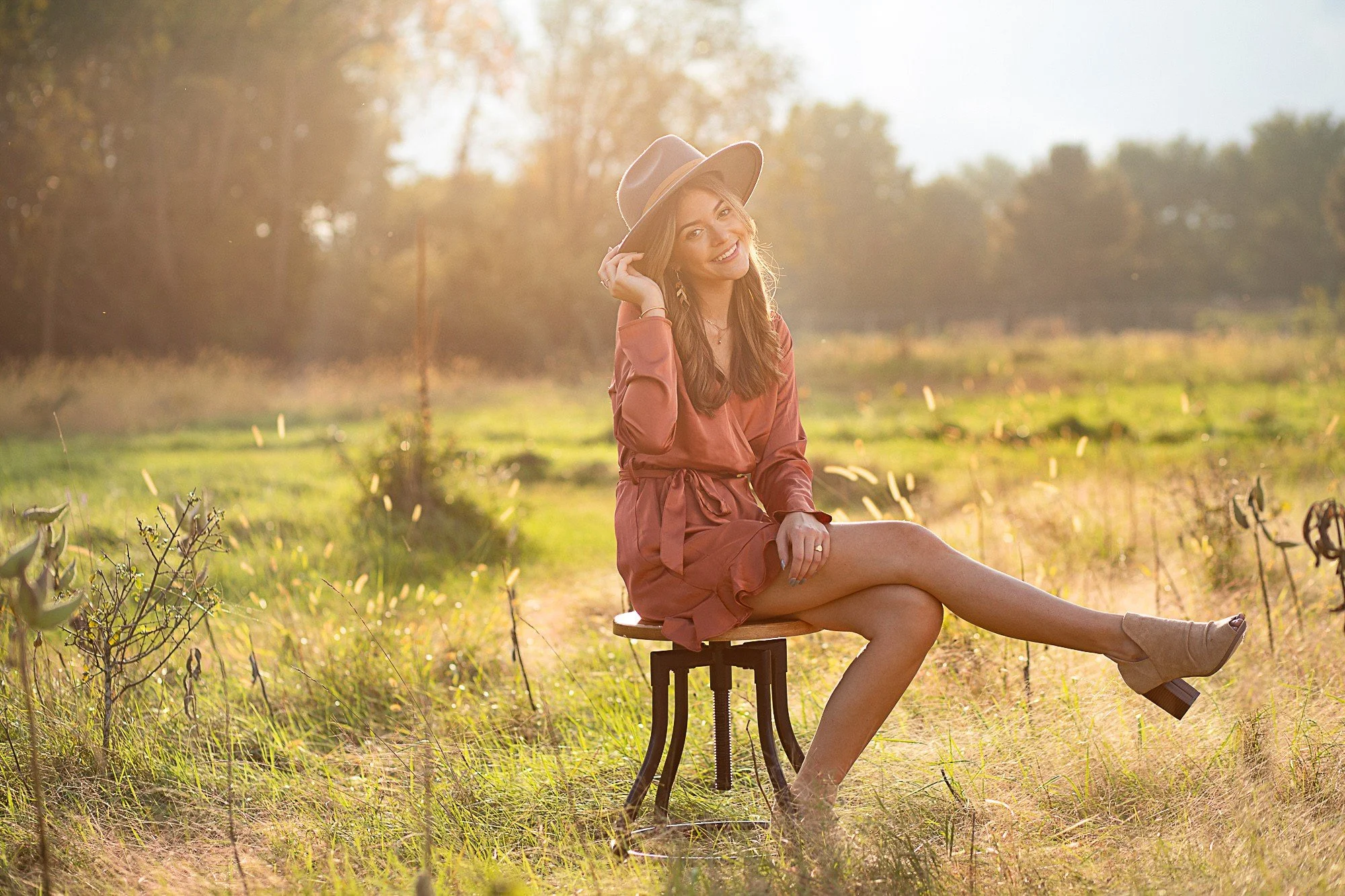 A woman sitting on a stool in a sunlit field, wearing a pink dress and a wide-brimmed hat, smiling and touching her hat with her right hand.
