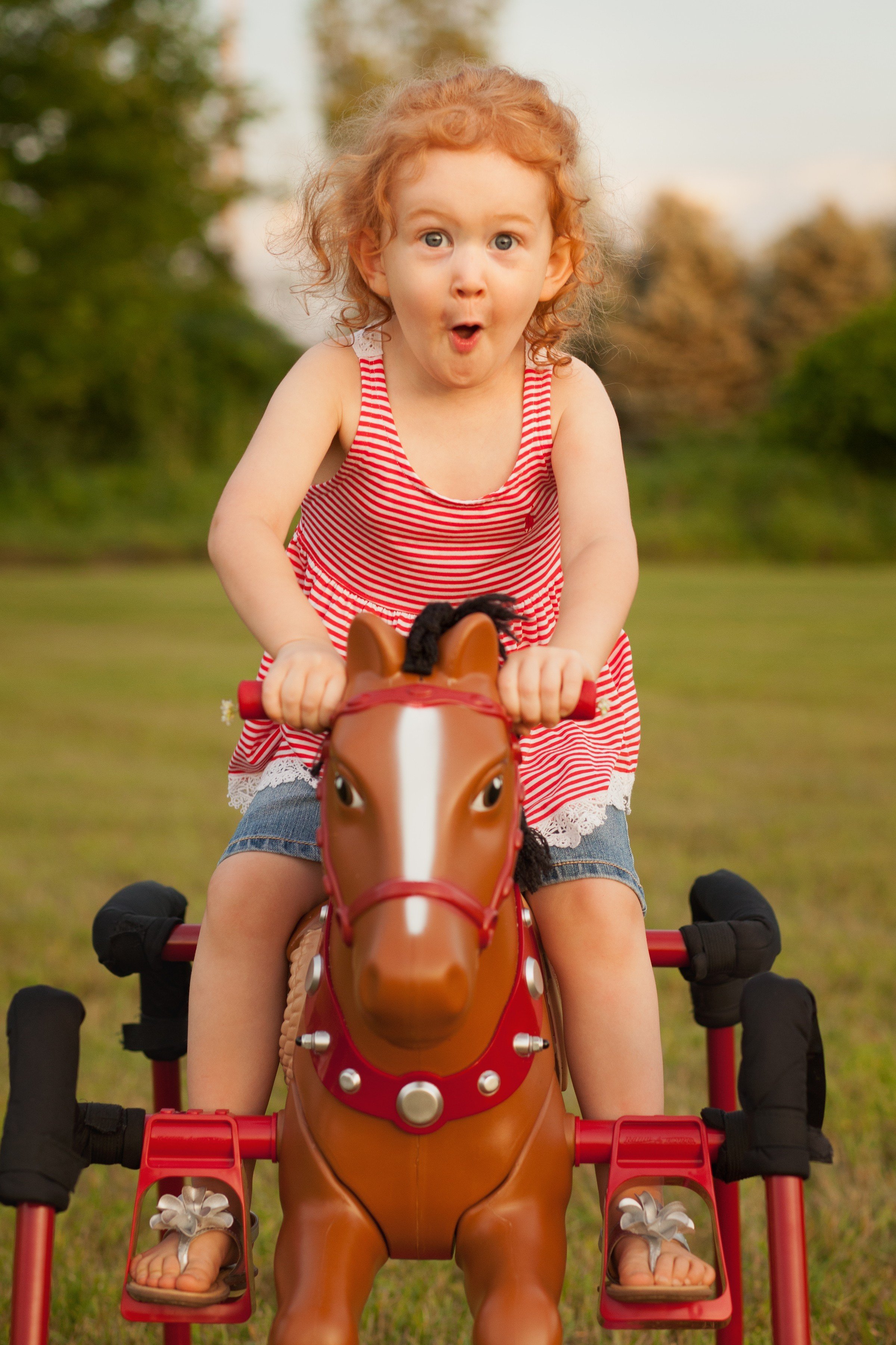 Young girl with curly red hair wearing a red and white striped dress, excitedly riding a brown plastic toy horse outdoors on green grass with trees in the background.
