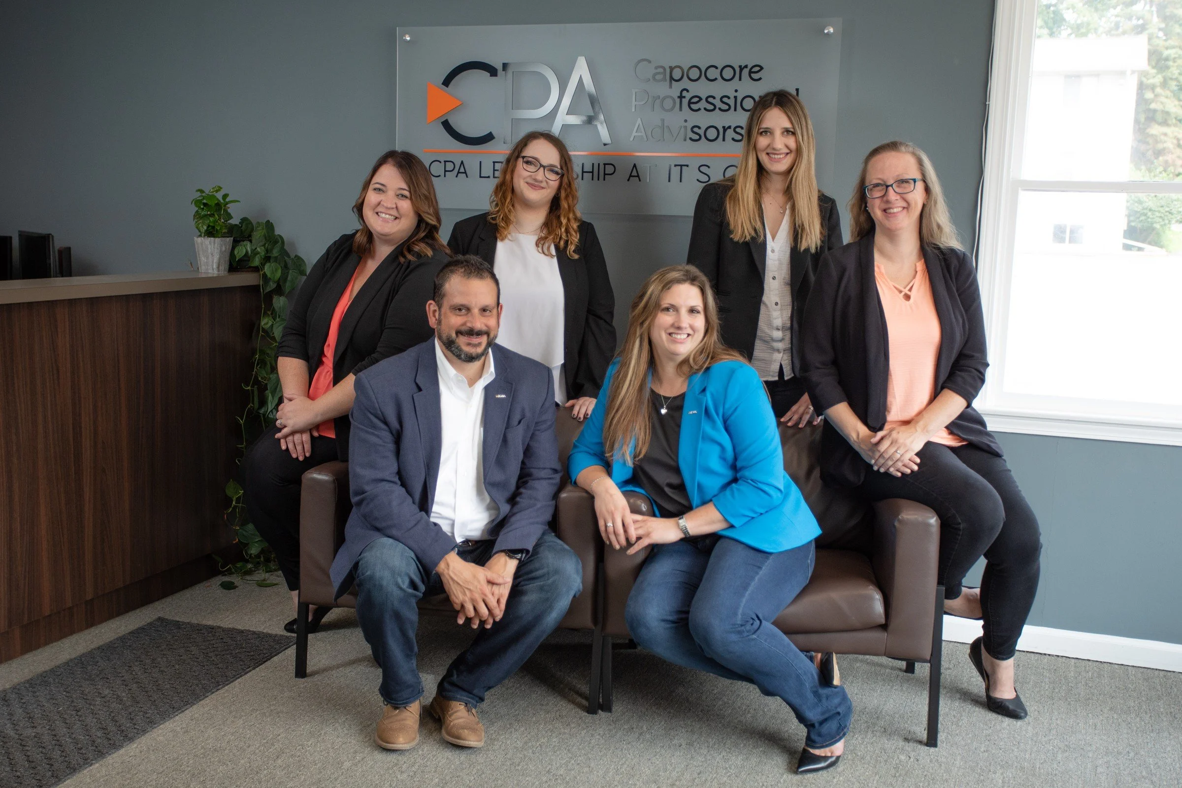 Group of seven professionals, five women and two men, posing indoors in front of a sign that says 'Capore Leadership Advisors' and 'CPA'. They are smiling and dressed in business casual and professional attire. One woman is seated on a brown loveseat