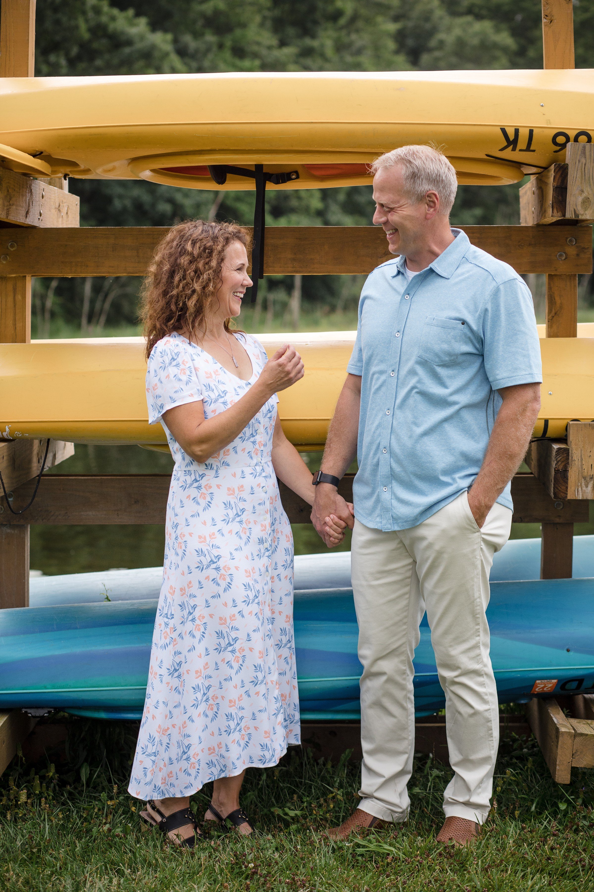 A smiling couple holding hands, standing outdoors near a rack of kayaks, with green trees in the background.
