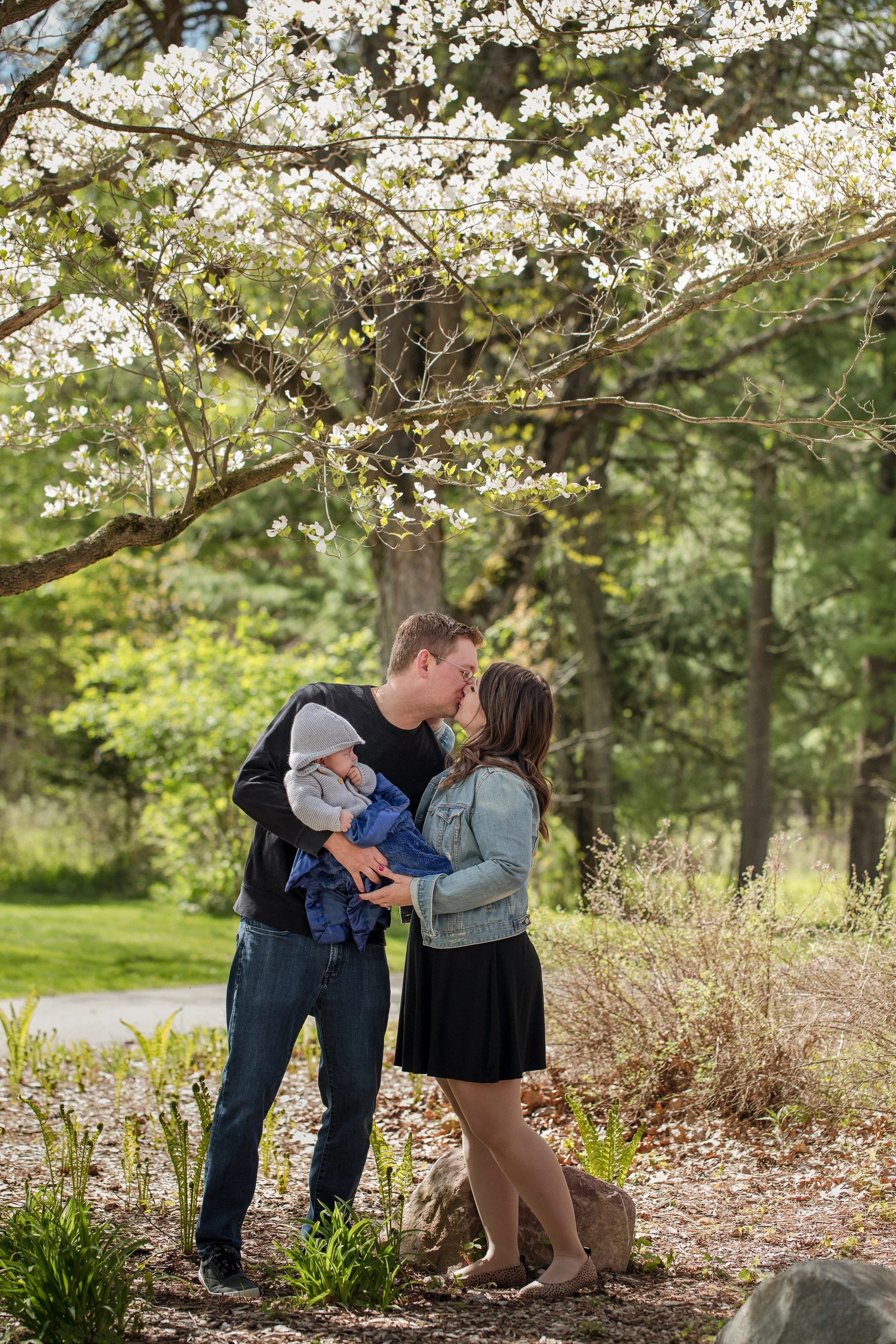 A family of three stands outdoors under flowering tree, sharing a kiss. The man holds a baby wrapped in a blanket, and the woman stands close to him, touching his arm. They are surrounded by greenery, rocks, and blossoming trees.