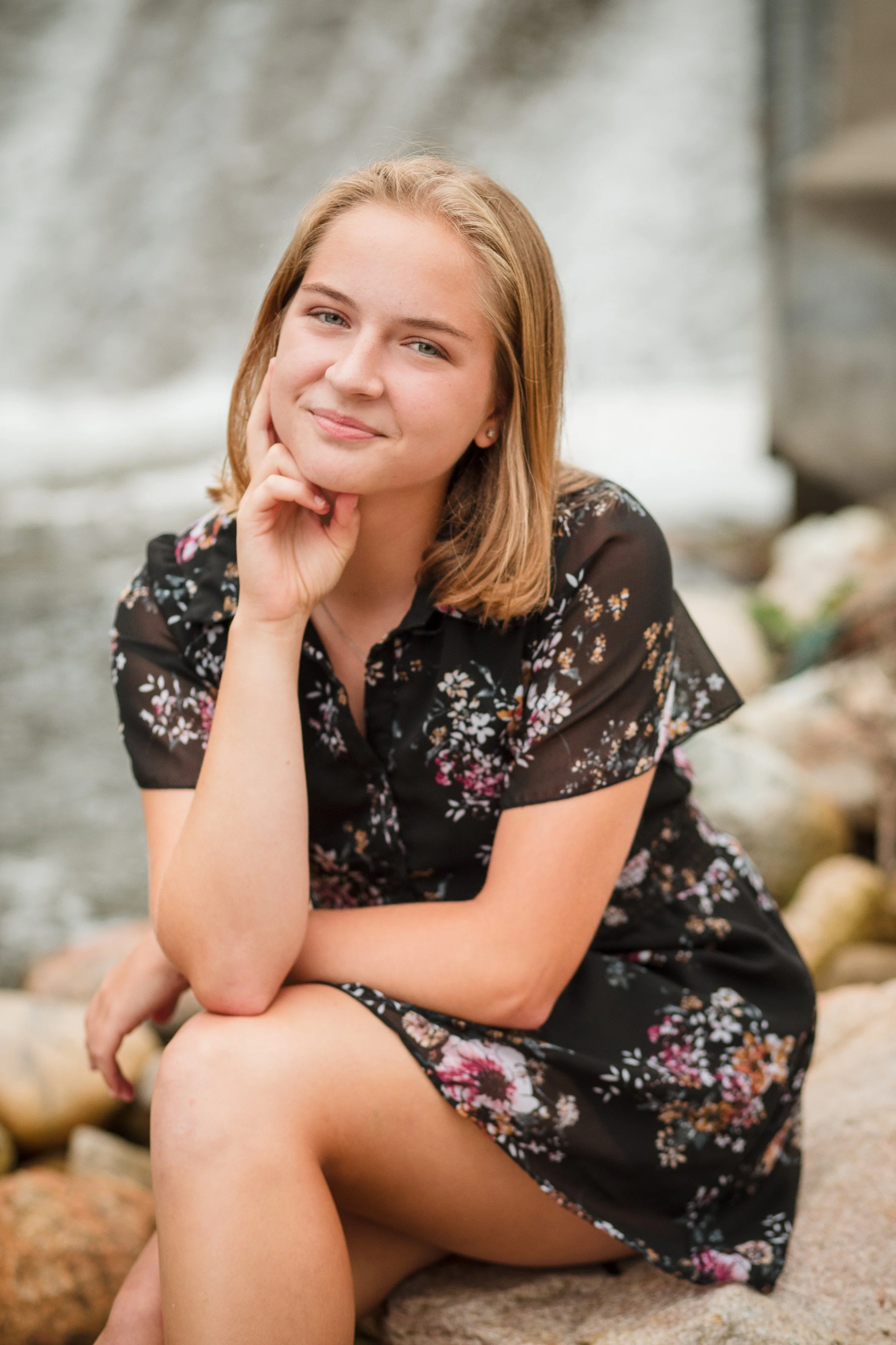 A young woman with shoulder-length brown hair sitting on rocks near a waterfall, wearing a black floral dress, smiling gently with her chin resting on her hand.