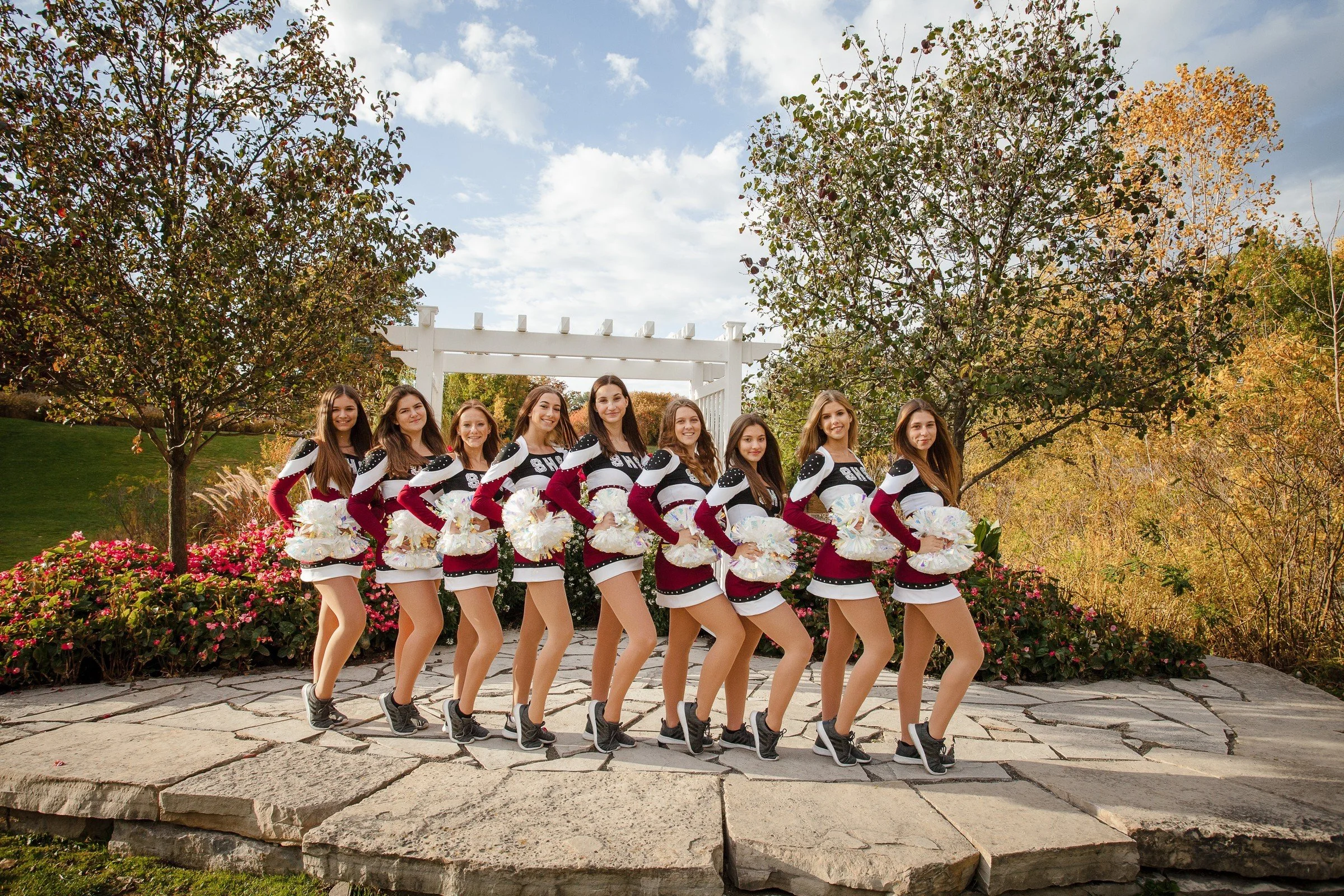 A group of cheerleaders dressed in maroon, black, and white uniforms holding pom-poms, standing outdoors on a stone pathway with trees and colorful autumn foliage in the background.