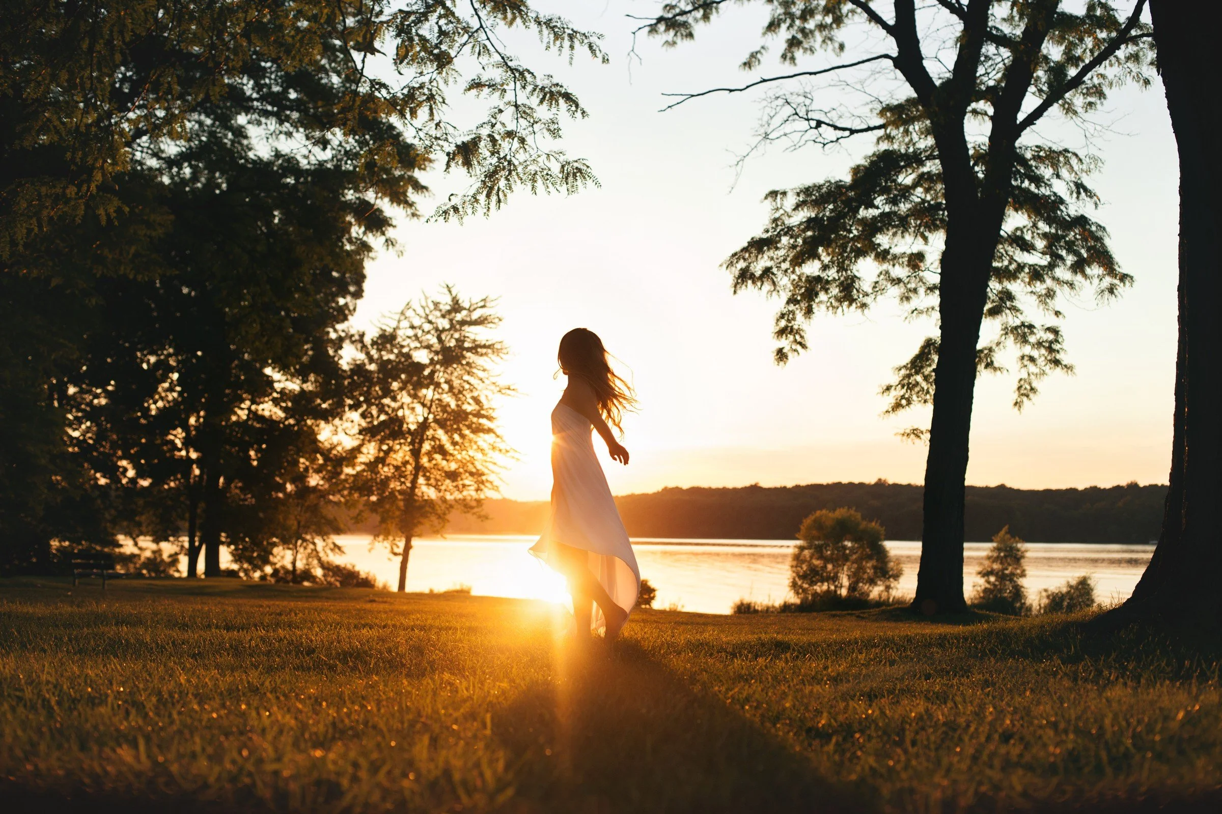 A woman in a white dress standing on grass near a river at sunset, with trees around her and the sun low in the sky.