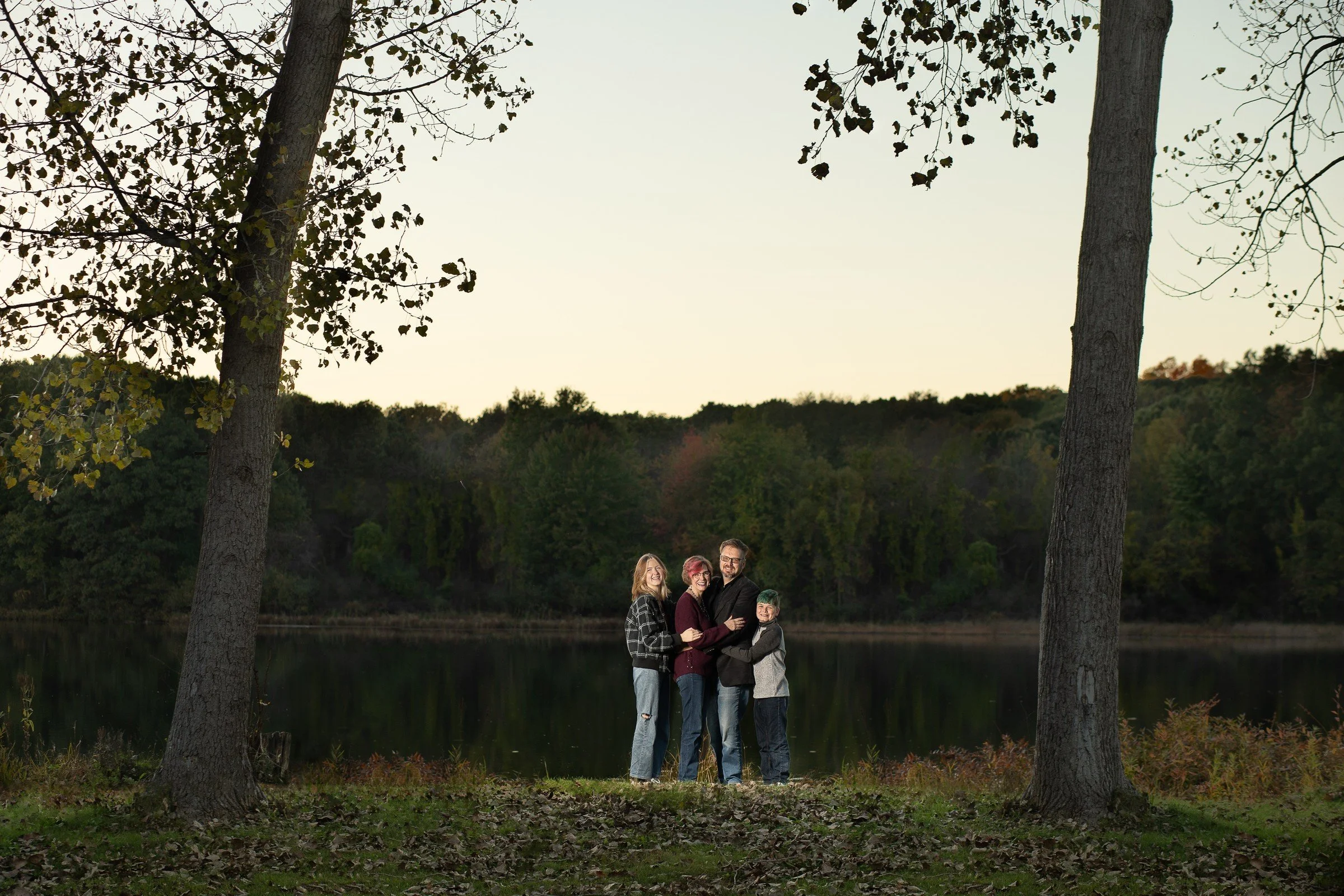 A family of four standing together near a lake in a wooded area during late afternoon or early evening.