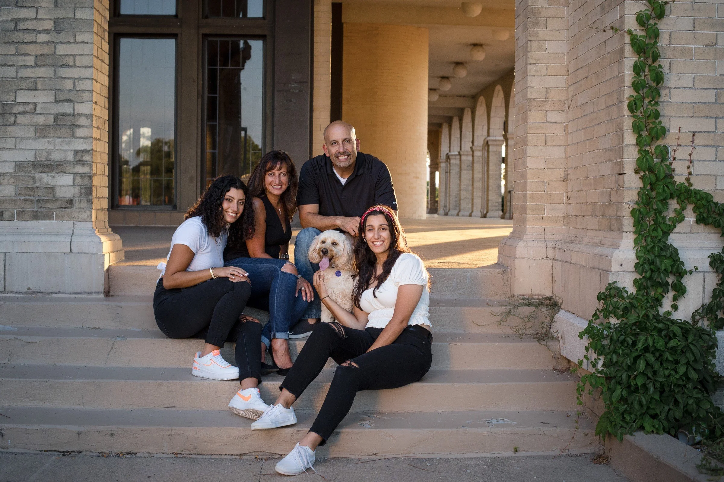 A family of four people and a dog sitting on steps outside a building during sunset, smiling at the camera.