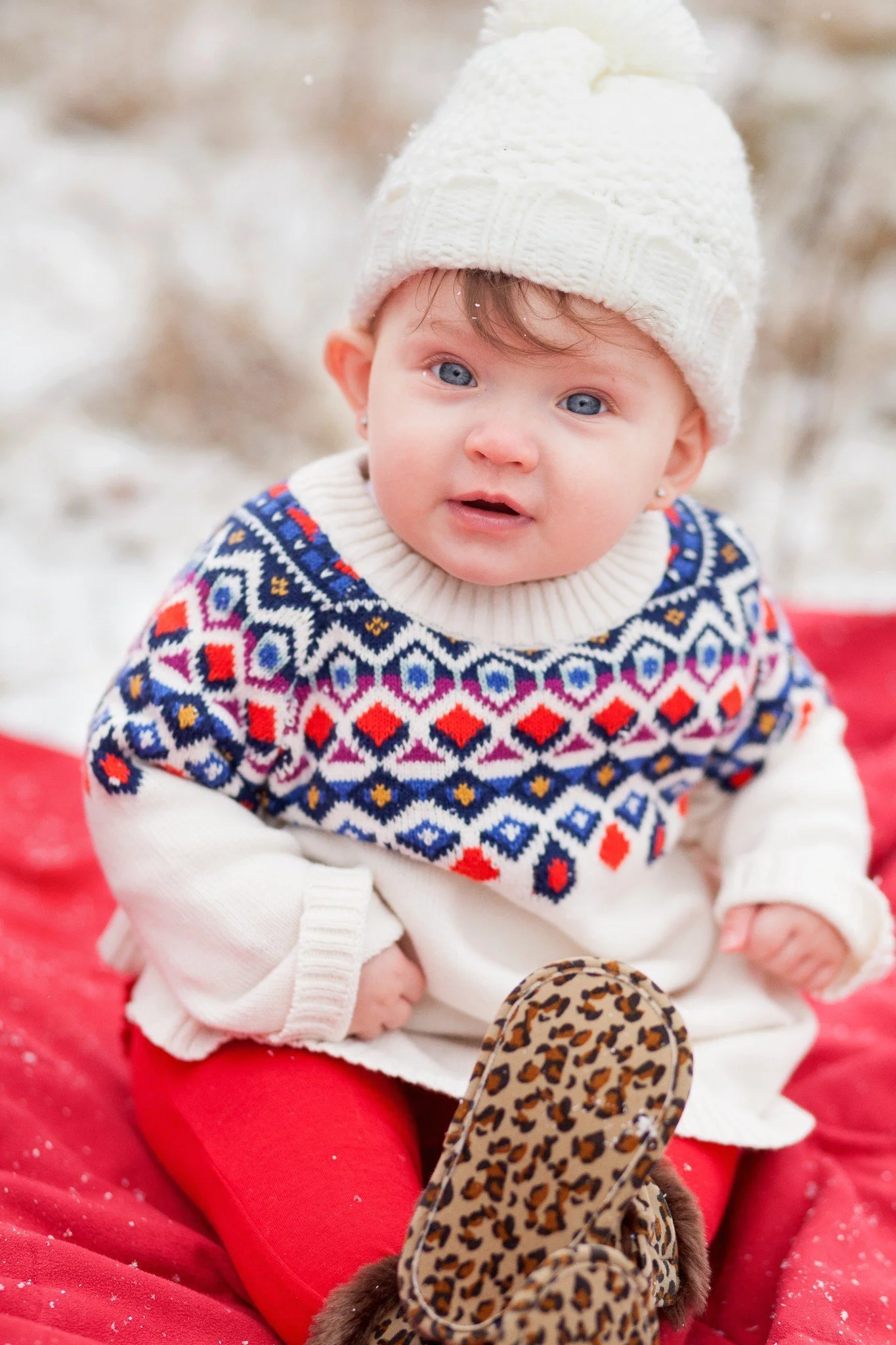 A baby girl with blue eyes wearing a white knit hat, a colorful patterned sweater, red pants, and leopard print shoes, sitting outdoors on a red blanket with a snowy background.