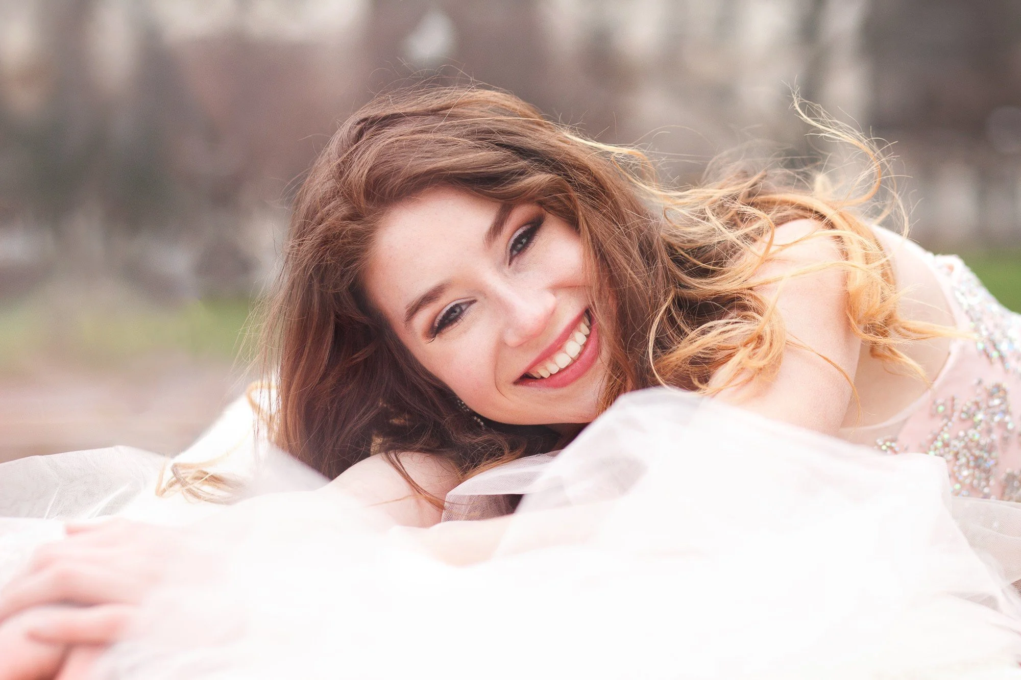 A woman with long, curly red hair laying outdoors on a soft surface, smiling at the camera, with a blurred background of trees.