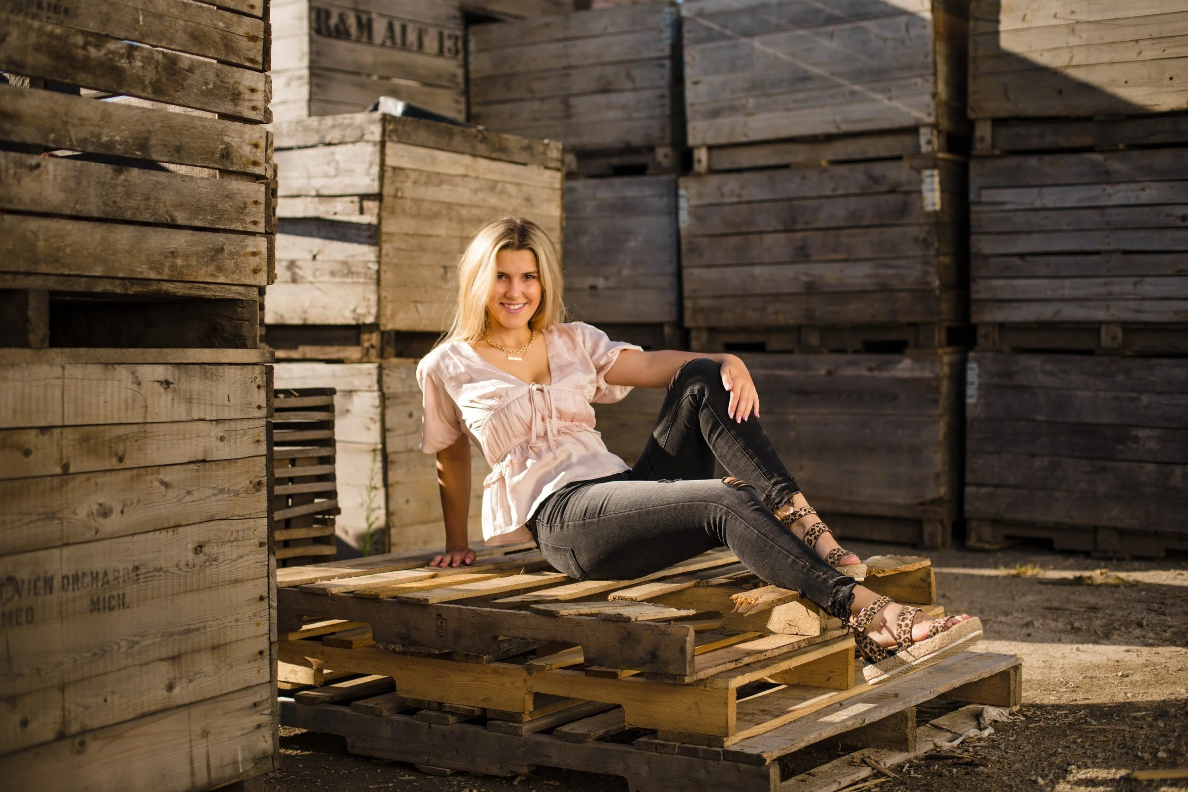 A young woman with blonde hair and a pink top sitting on stacked wooden pallets among large wooden crates, smiling at the camera.