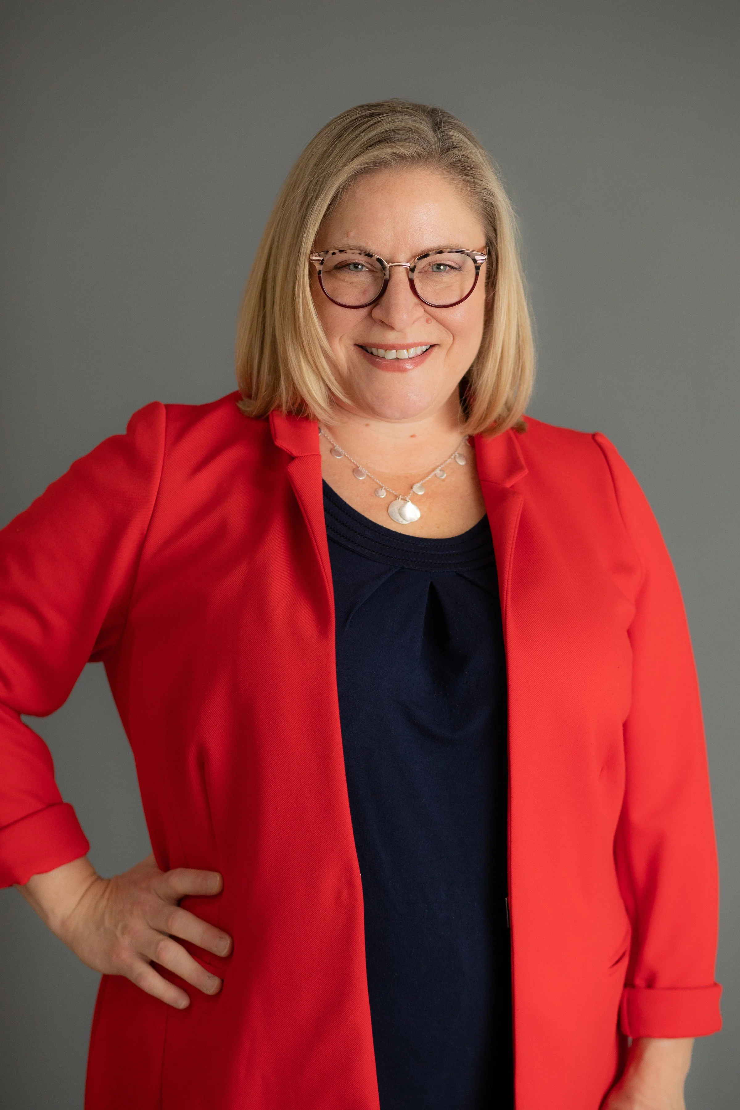 A woman with blonde hair, wearing glasses, a navy blue top, a red blazer, and a silver necklace, smiling with her hand on her hip against a gray background.
