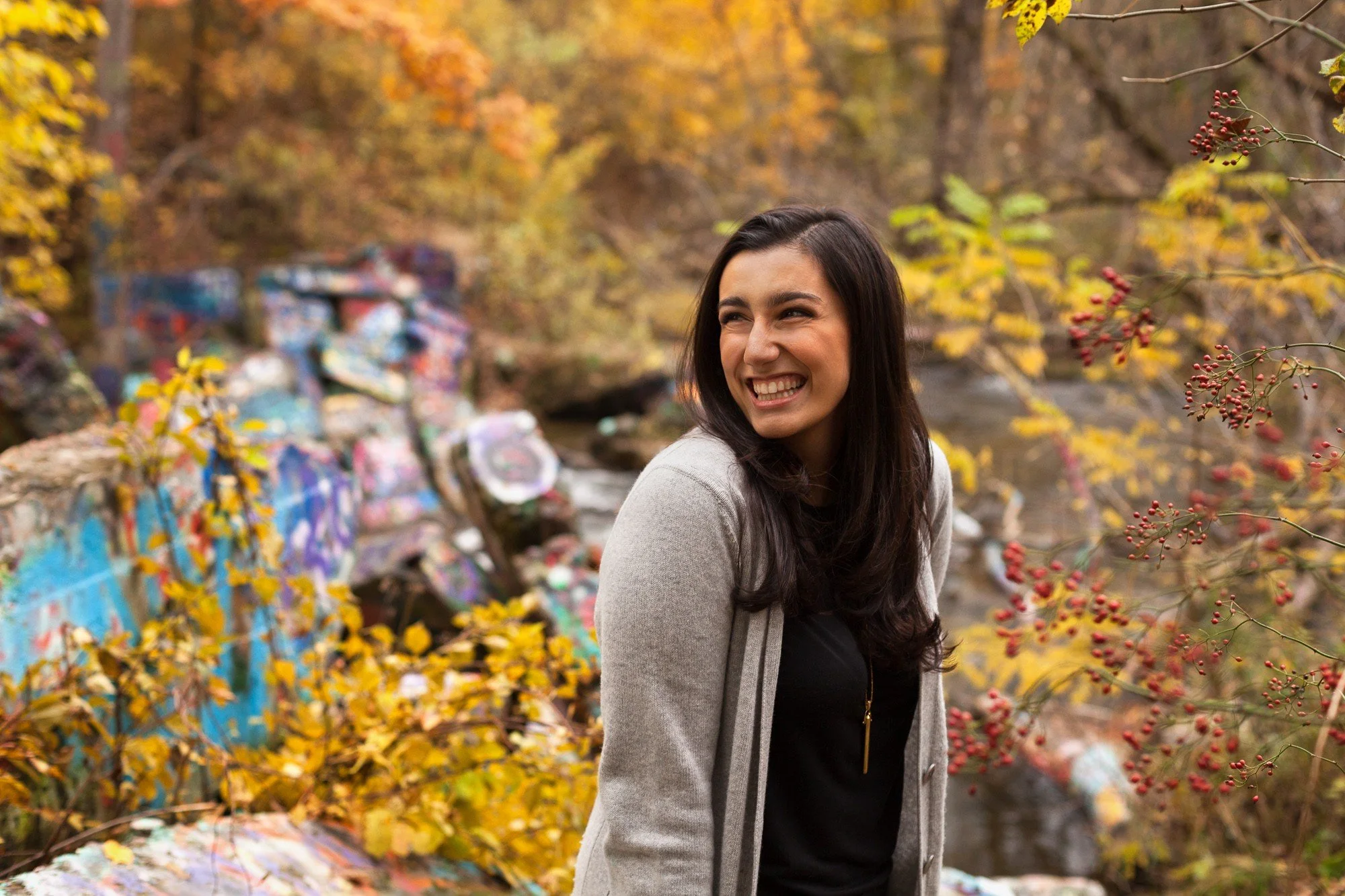 A young woman with dark hair smiling outdoors in autumn, standing near a river with colorful graffiti on rocks and yellow and orange fall foliage in the background.