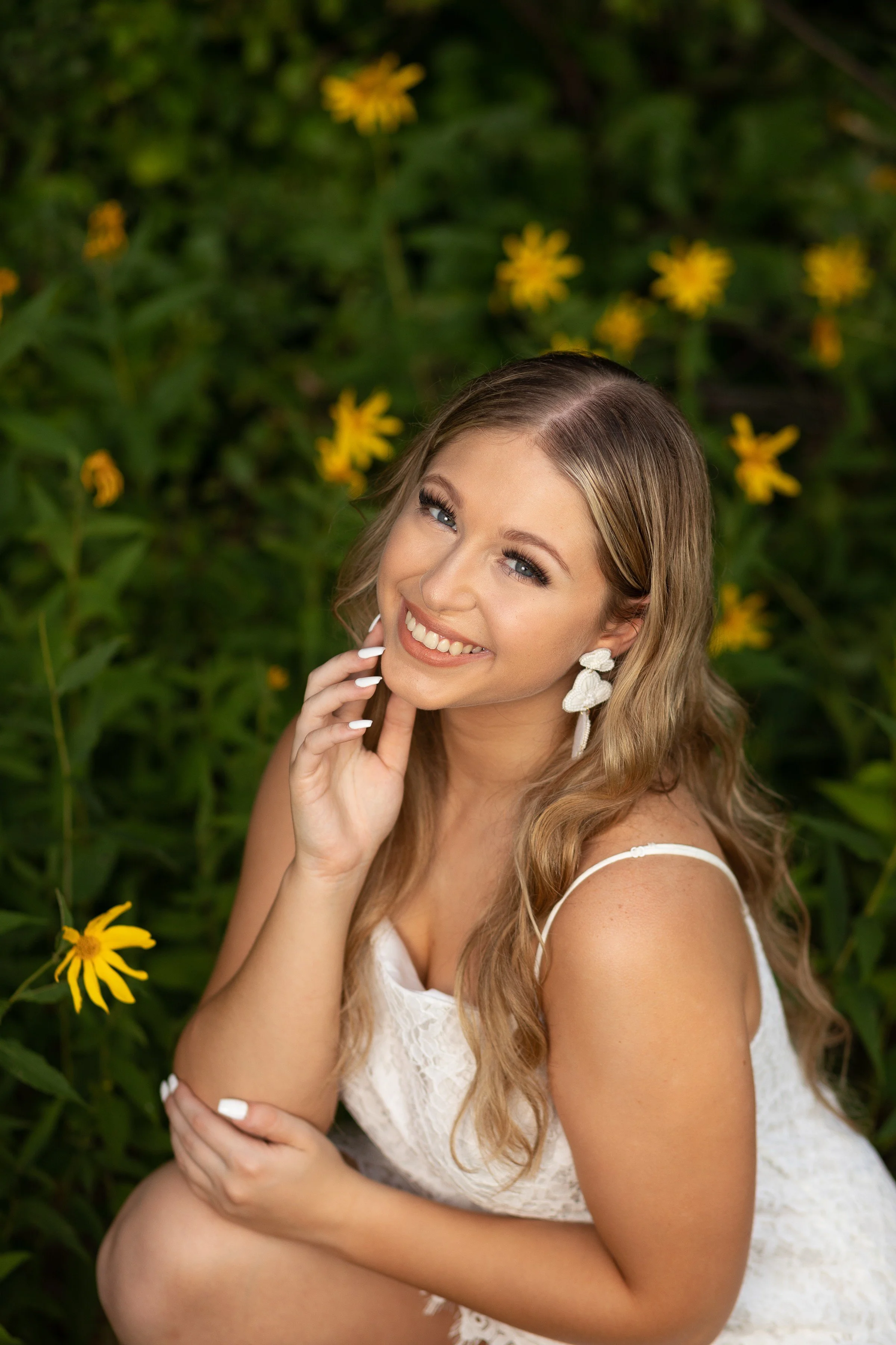 A woman with long wavy hair, smiling, wearing butterfly earrings and a white lace dress, sitting among yellow flowers in a garden.