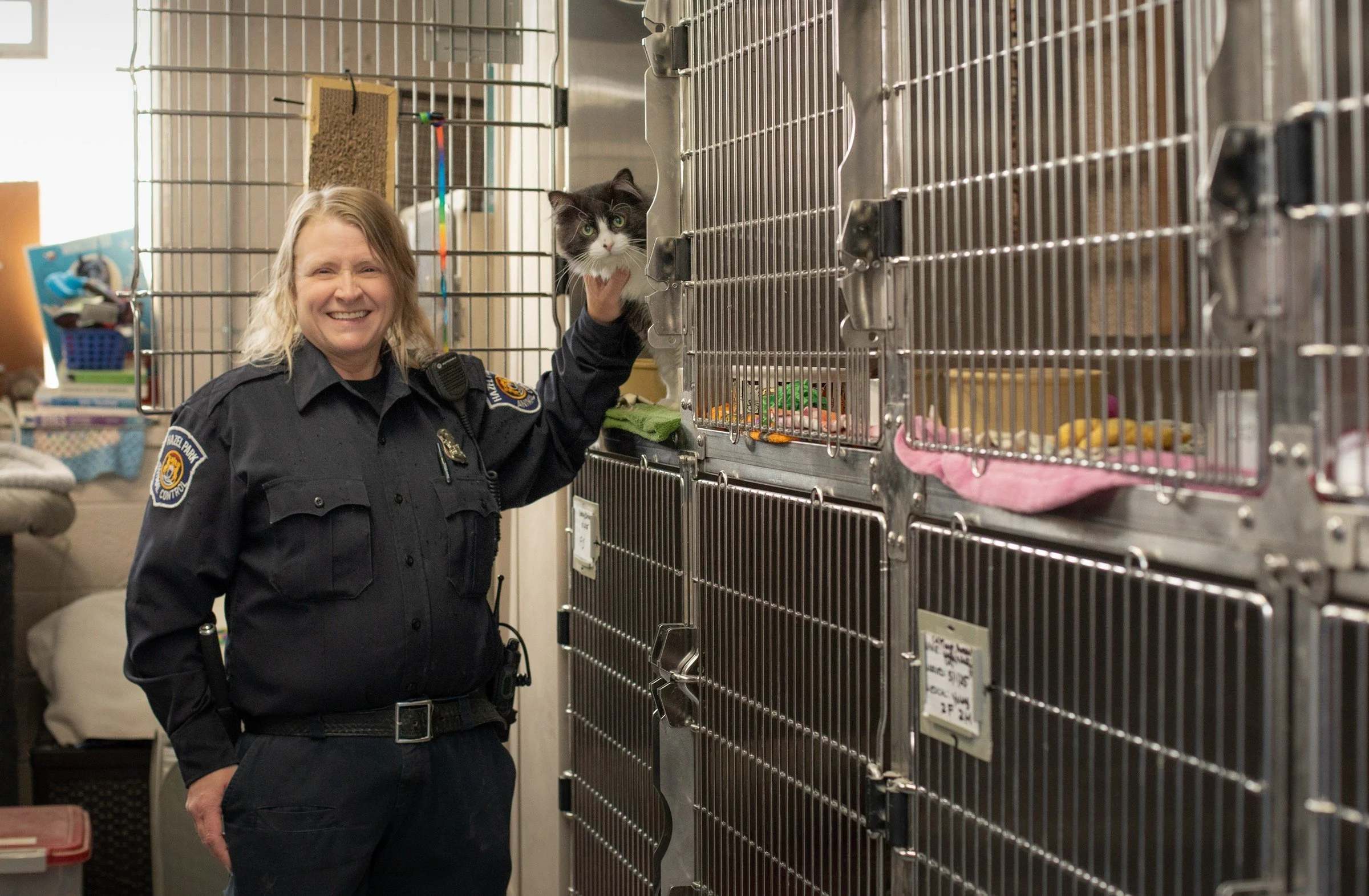A woman in a police uniform smiling and holding a black and white cat in a shelter with metal cages.