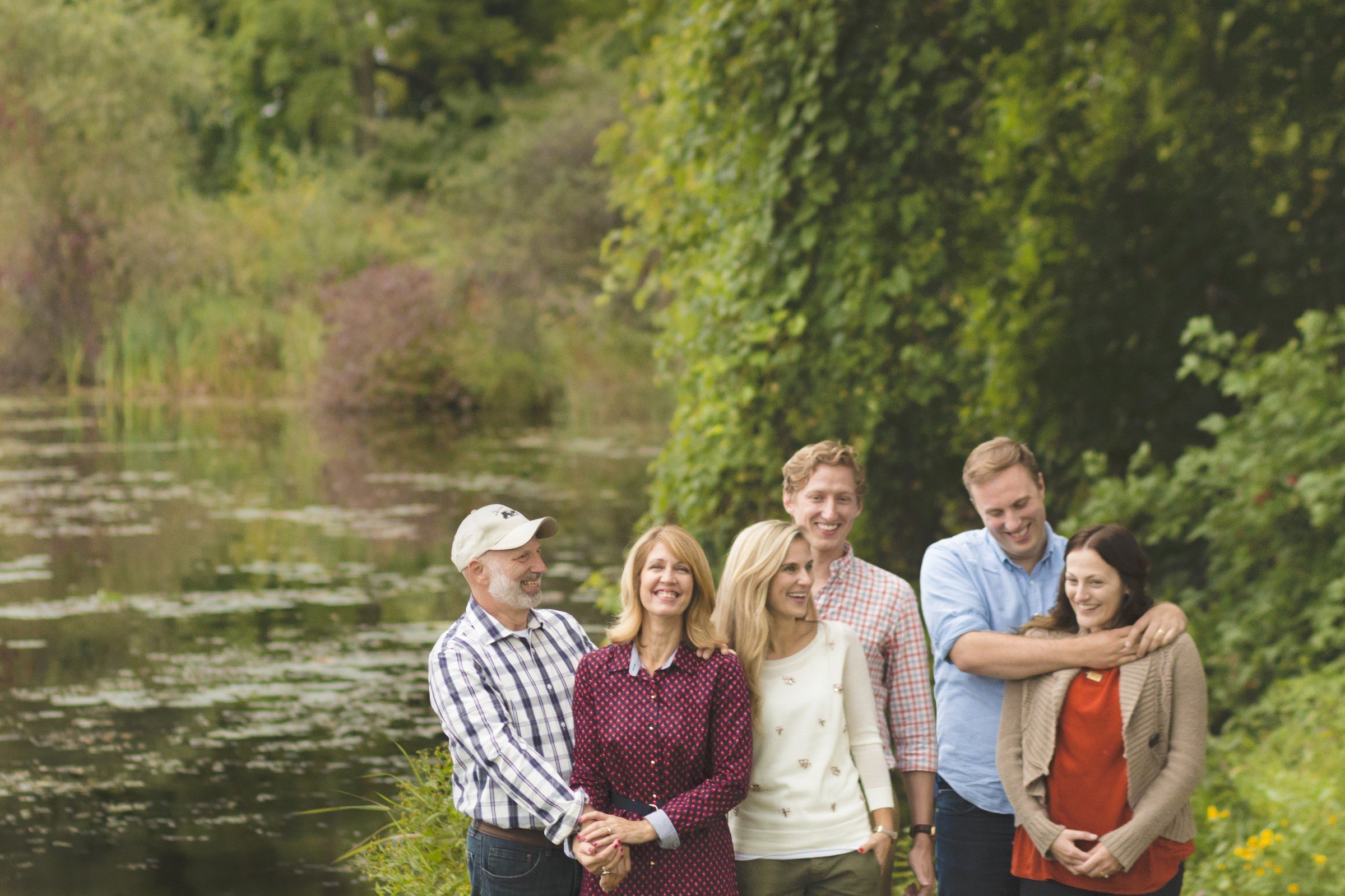 Six people standing outdoors near a lake or river, smiling and enjoying nature, surrounded by trees and greenery.