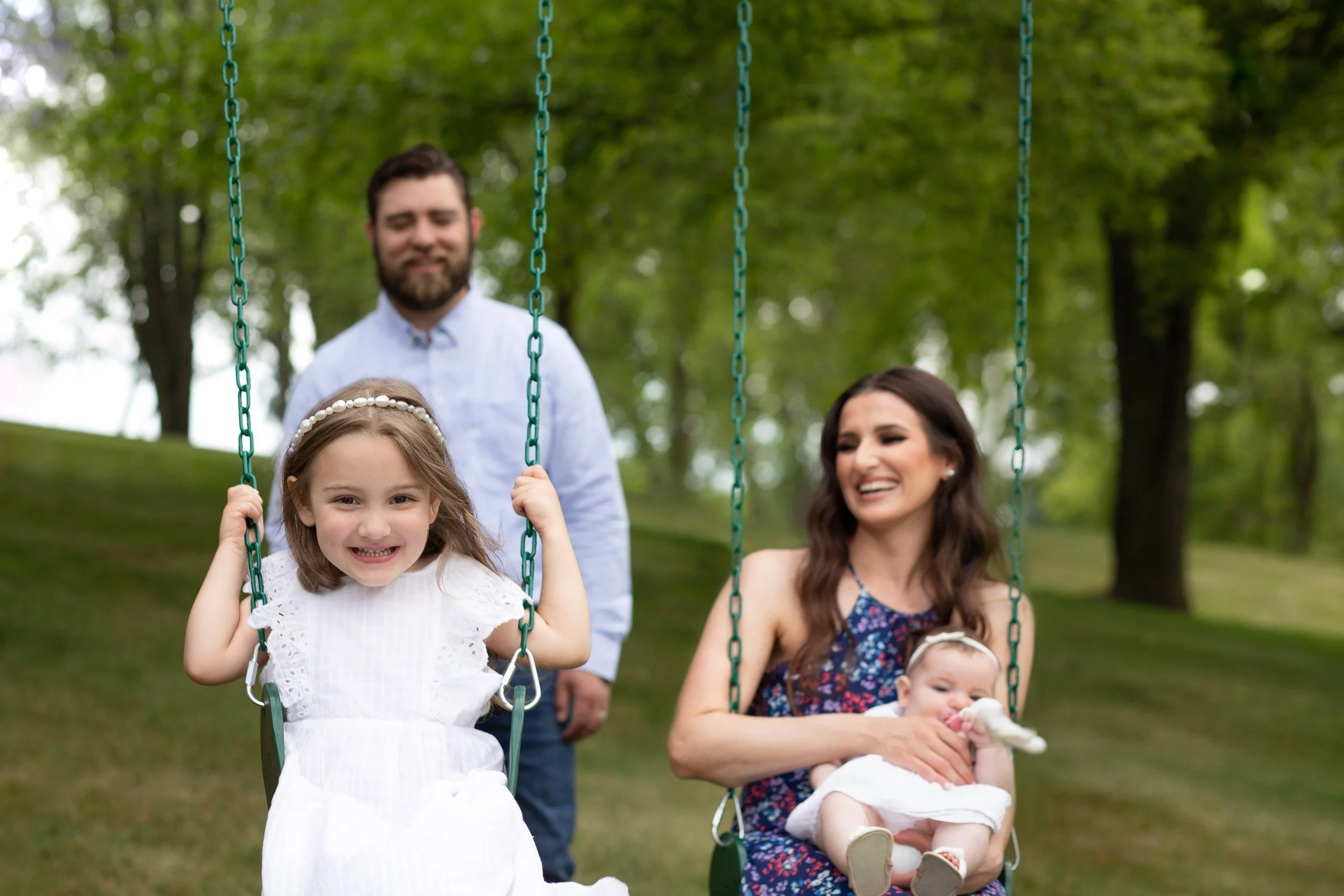 A family in a park with a girl on a swing, a woman holding a baby on another swing, and a man standing nearby behind them, surrounded by green trees.