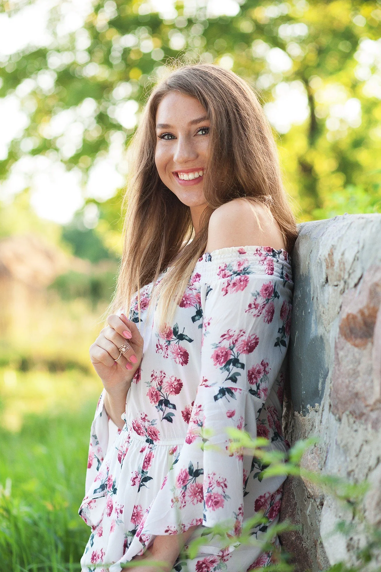 Young woman with long, light brown hair, smiling, wearing a white floral off-shoulder dress, leaning against a stone wall outdoors with green trees and sunlight in the background.