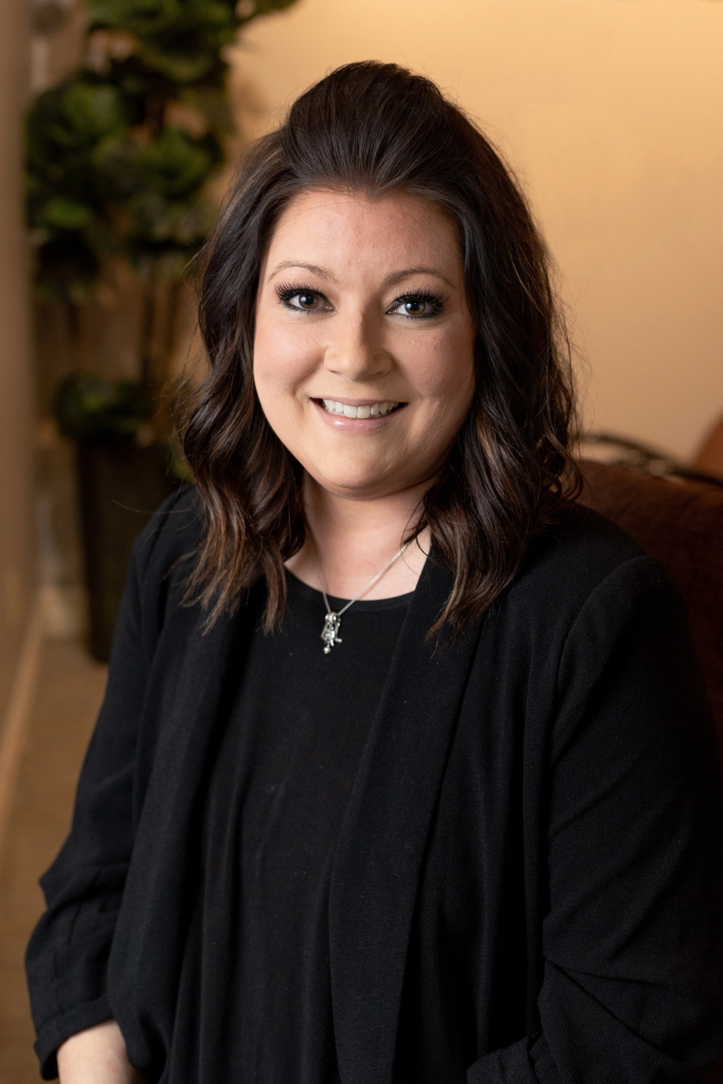 A woman with shoulder-length wavy dark brown hair, wearing a black top and jacket, smiling at the camera, sitting indoors with a blurred background and a plant in the background.
