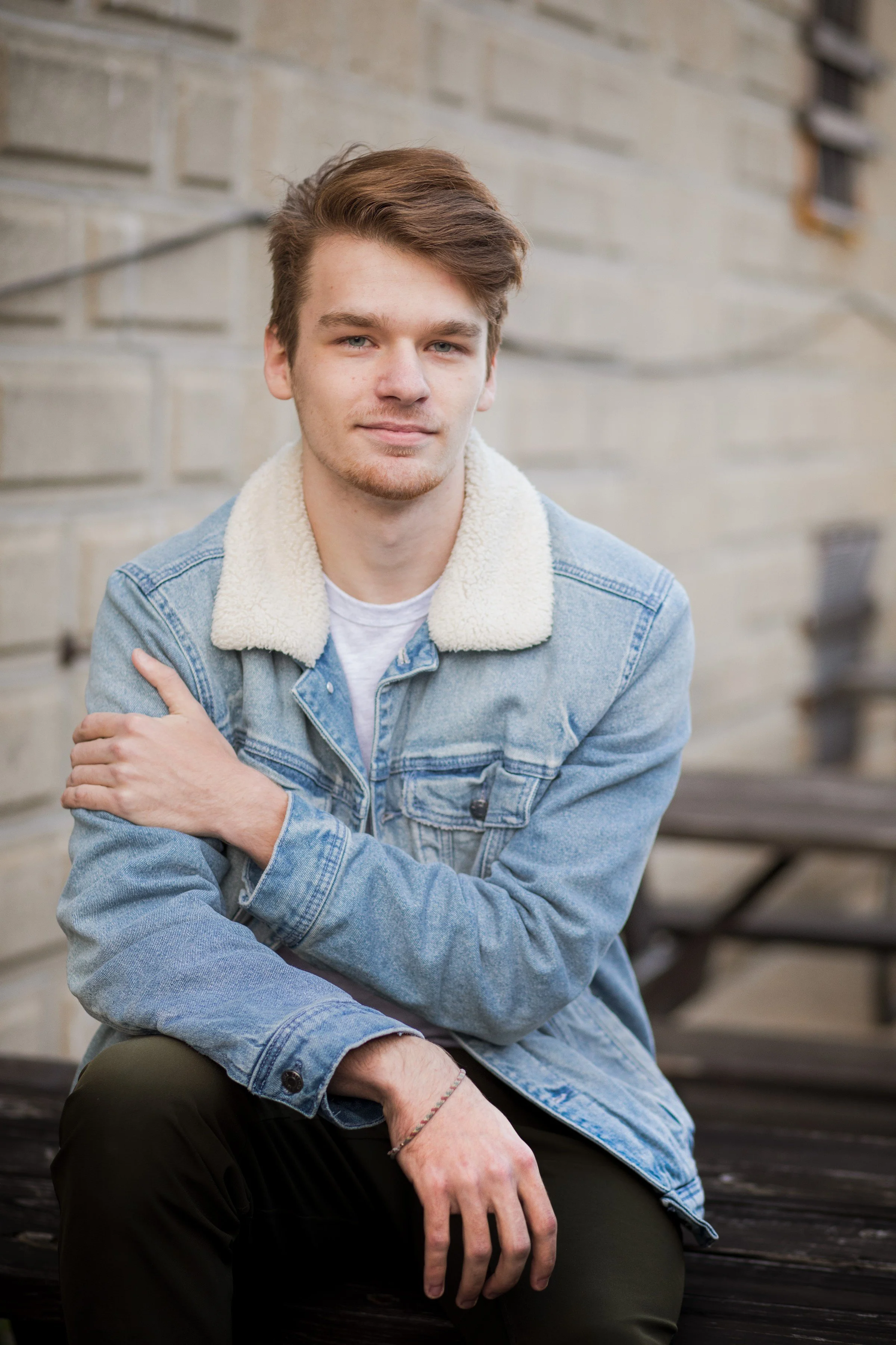 A young man with brown hair, blue eyes, and light facial hair, sitting outdoors on a wooden bench. He is wearing a light blue denim jacket with a white shearling collar and a white T-shirt underneath, with a neutral expression and a slight smile.