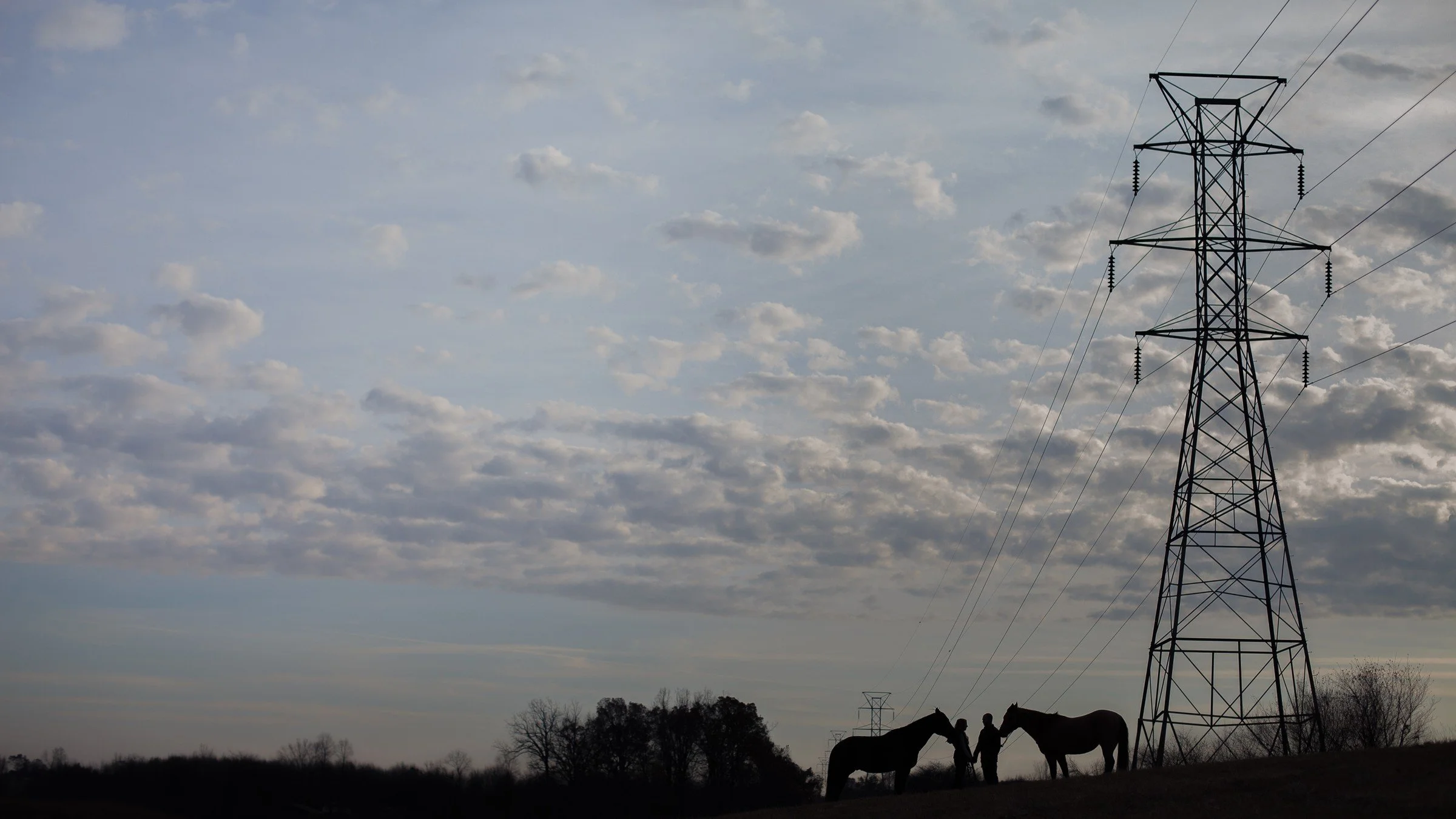 Silhouettes of two horses and two people standing near a large electrical tower against a cloudy sky.