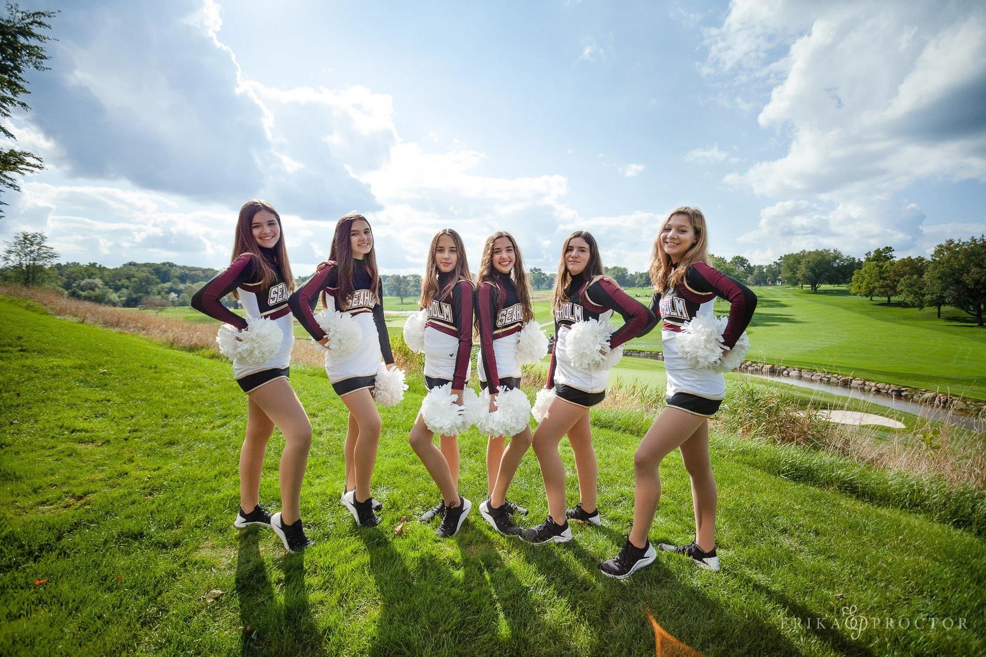Six cheerleaders in maroon and white uniforms with pom-poms standing outdoors on lush green grass, with a golf course and trees in the background under a partly cloudy sky.