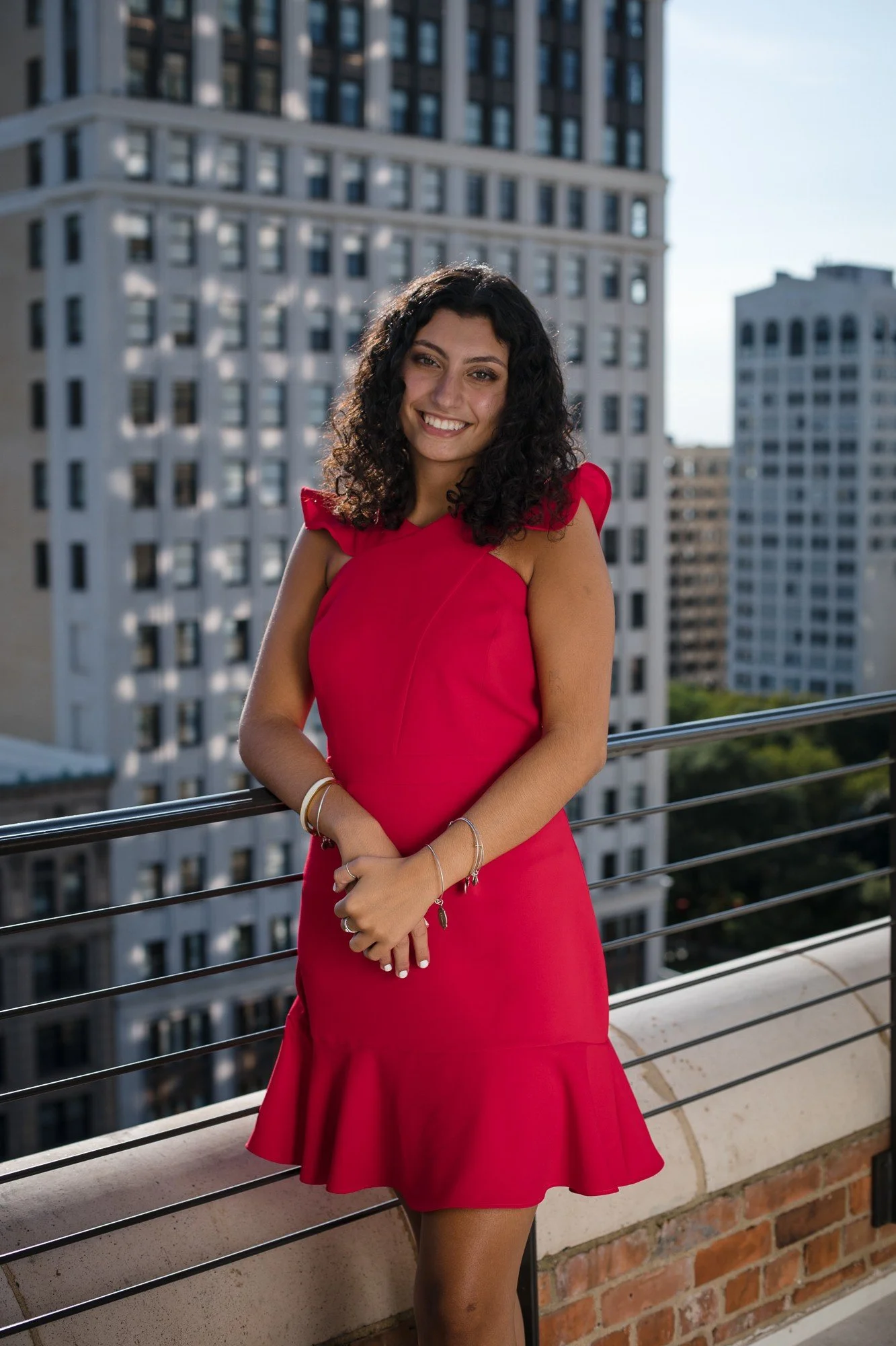 A woman in a red dress smiling on a balcony with city buildings in the background.