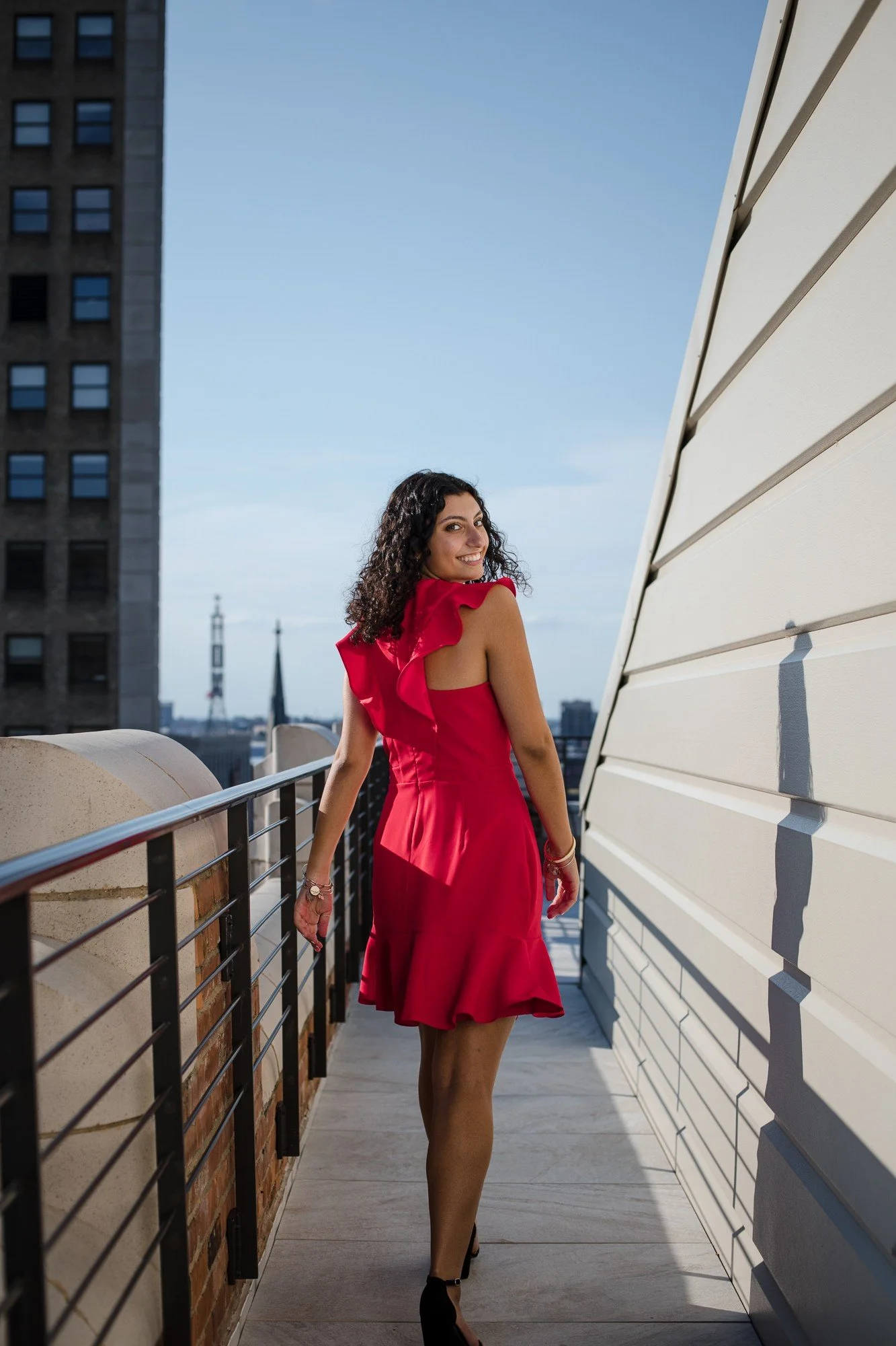 A woman in a red dress walking on a rooftop balcony in an urban area, with a cityscape and buildings in the background during daytime.