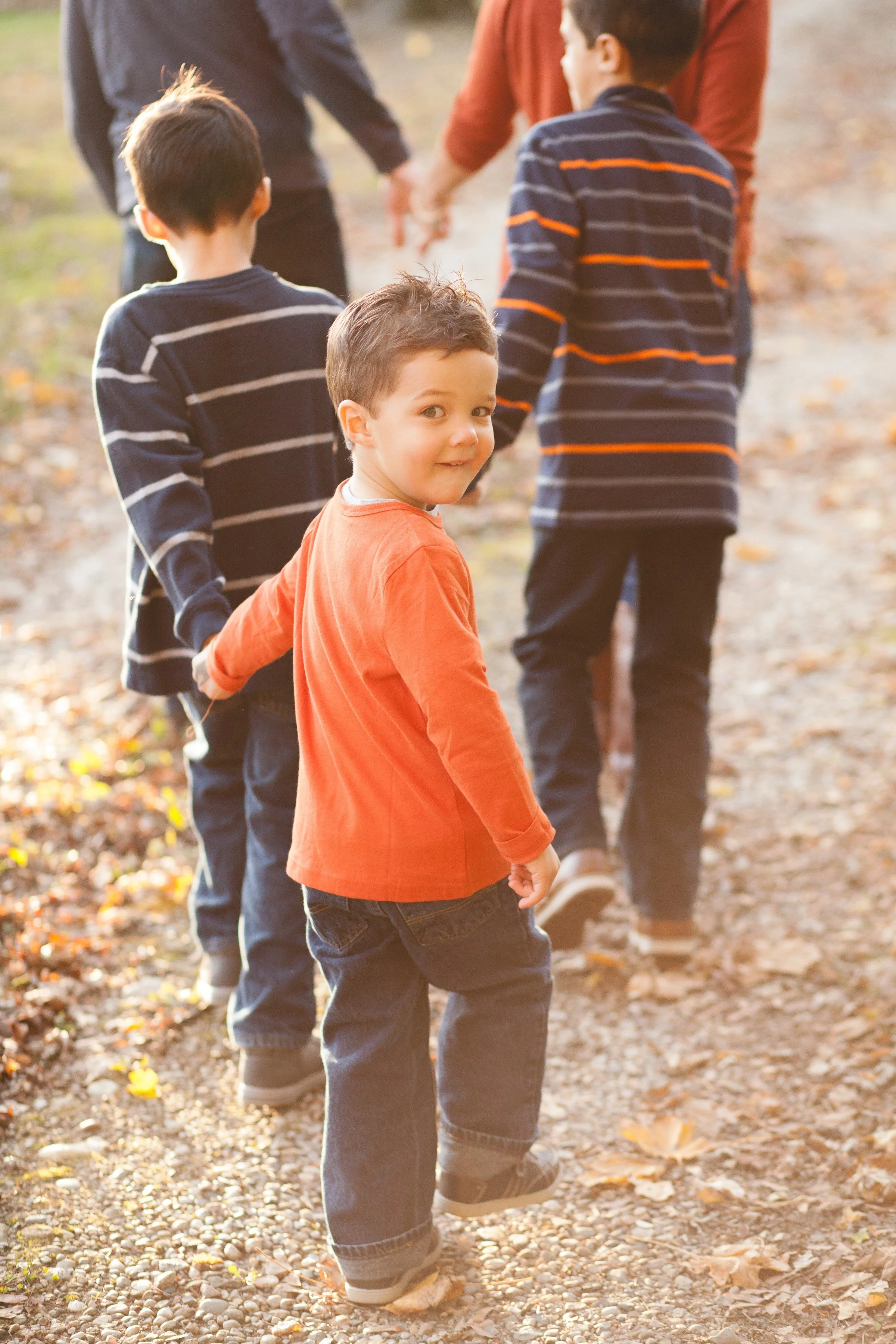 A young boy with brown hair in an orange shirt and jeans holding hands with other kids and adults while walking along a tree-lined path covered in fallen leaves.