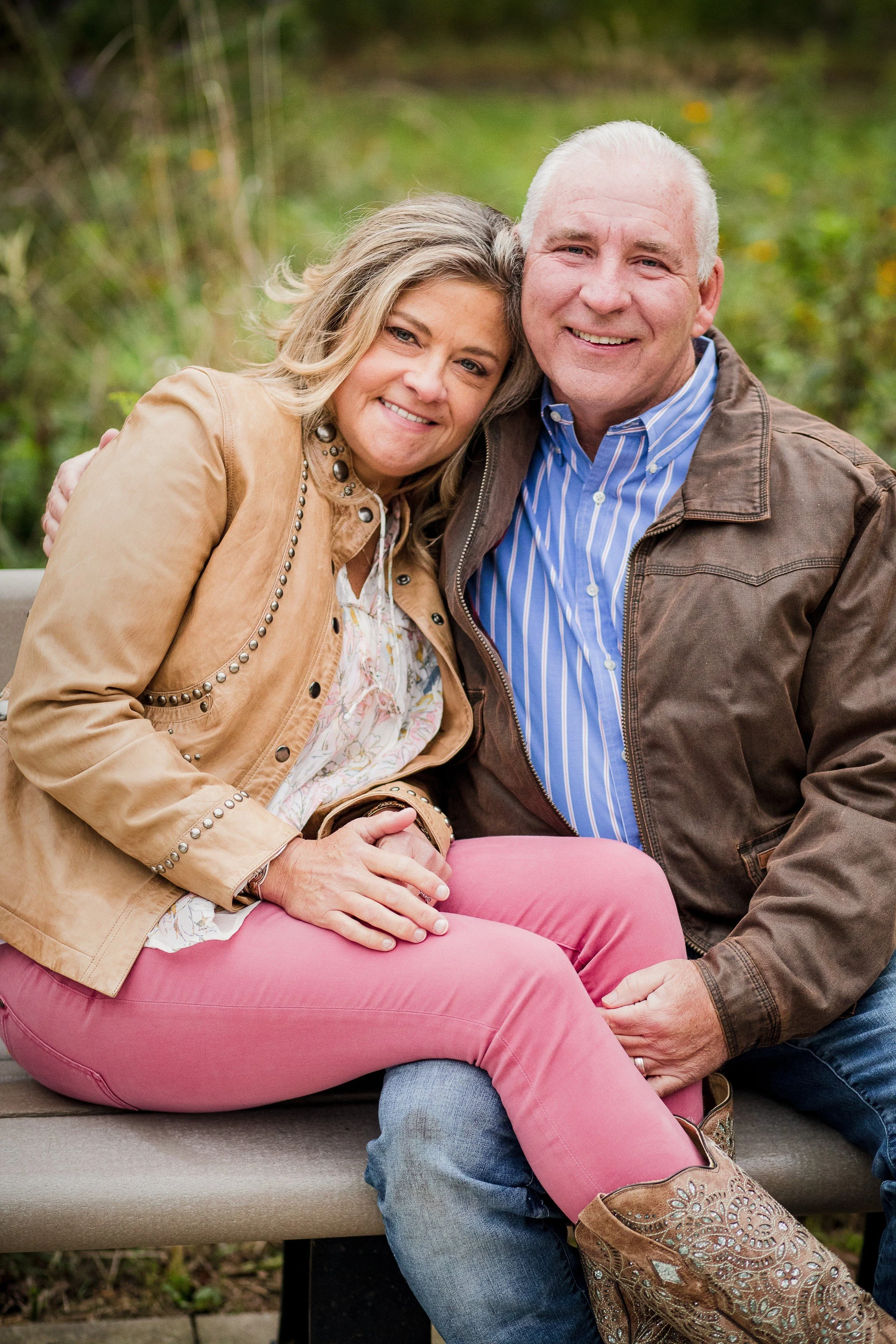 A smiling middle-aged couple sitting close together outdoors on a park bench, embracing each other. The woman has blonde hair and is wearing a tan jacket, a white patterned shirt, and pink pants. The man has white hair and is wearing a brown jacket o